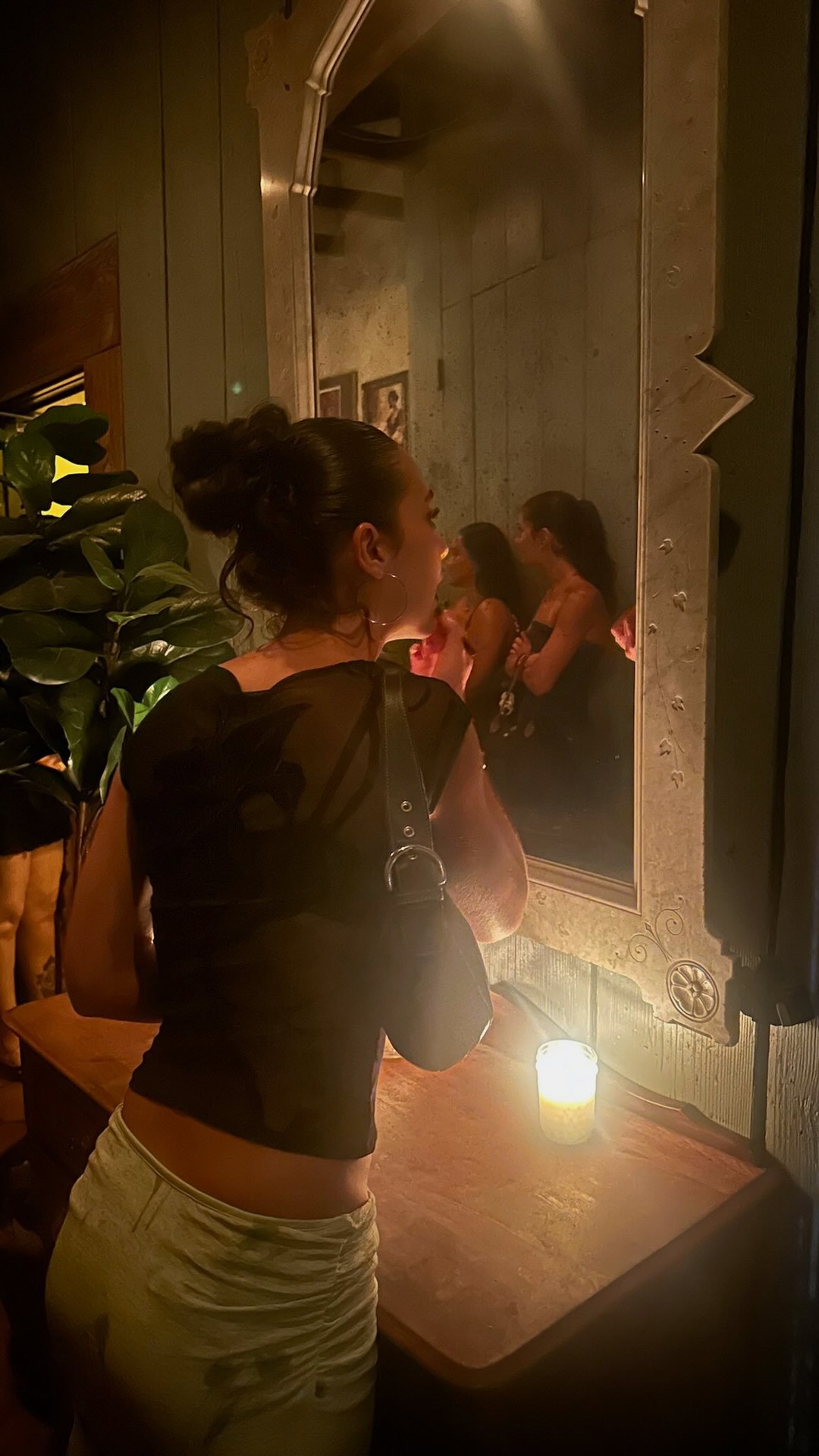 A young woman with her hair in a bun is applying lipstick in front of a mirror in a dimly lit room. She is wearing a sheer black top and light-colored ruched pants. In the reflection of the mirror, another woman is visible, also applying makeup. A lit candle sits on the wooden vanity in front of the mirror. The text "holding on to every last bit of summer with these pieces from @princesspolly use code GISELE20 !!" is overlaid on the image.