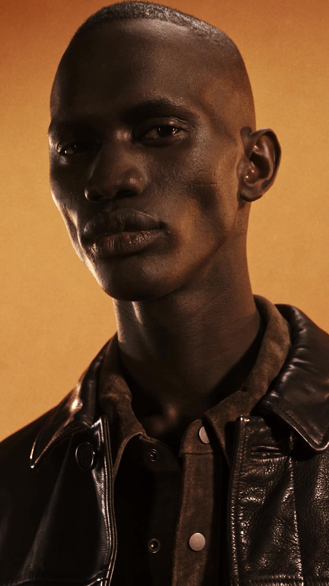 A close-up, studio portrait of a dark-skinned man with a shaved head, looking directly at the camera. He is wearing a dark leather jacket over a brown suede shirt. The background is a warm, textured orange. Text overlaying the image asks, "Ready to turn up the volume on your looks?" The copy continues, "Our leathers are iconic – find your new forever favourite."