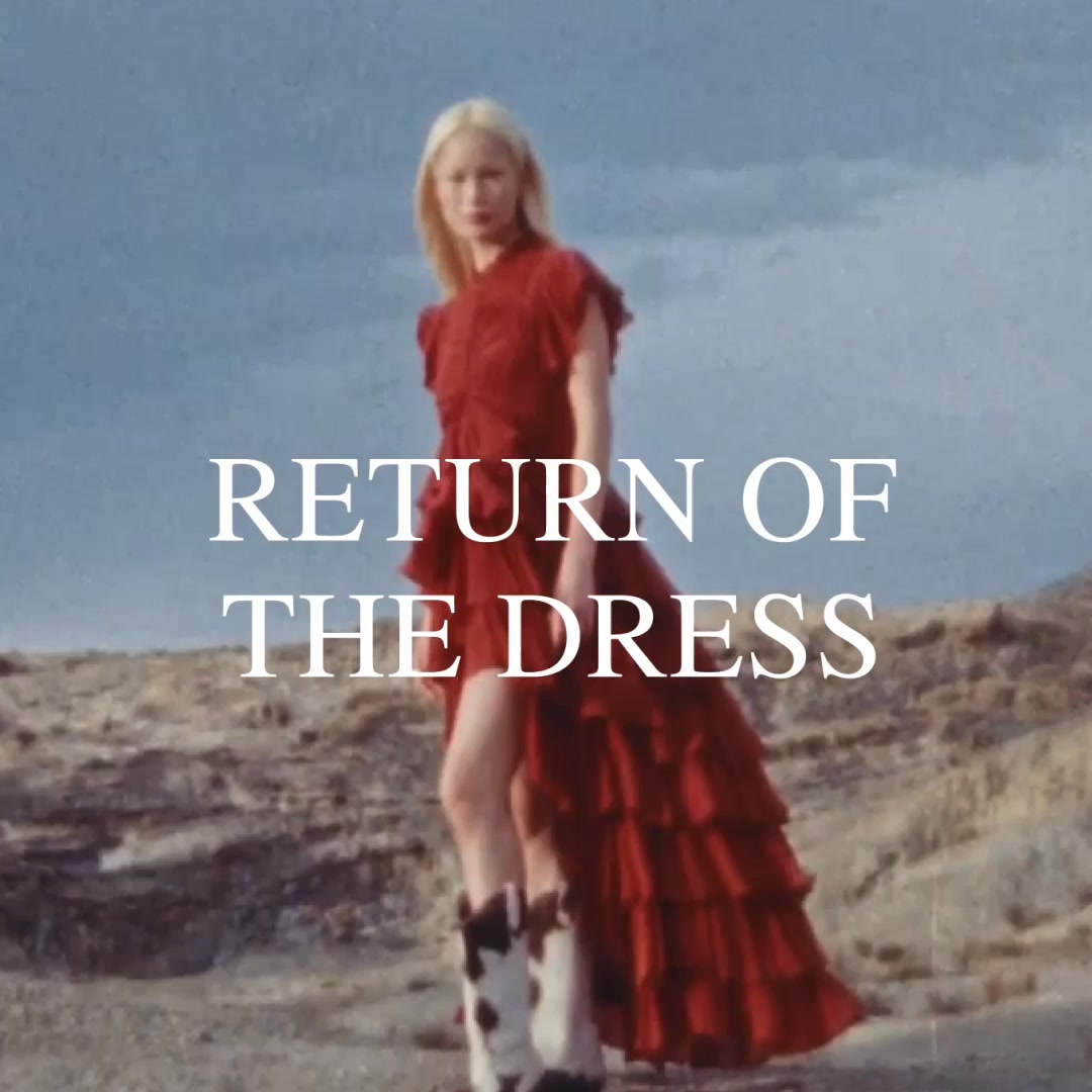 A woman with blonde hair, wearing a long, flowing red dress and cowboy boots, stands in a barren, outdoor landscape under a cloudy sky. Large white text overlaid on the image reads "RETURN OF THE DRESS." The overall aesthetic is dramatic and slightly windswept, suggesting a fashion editorial.