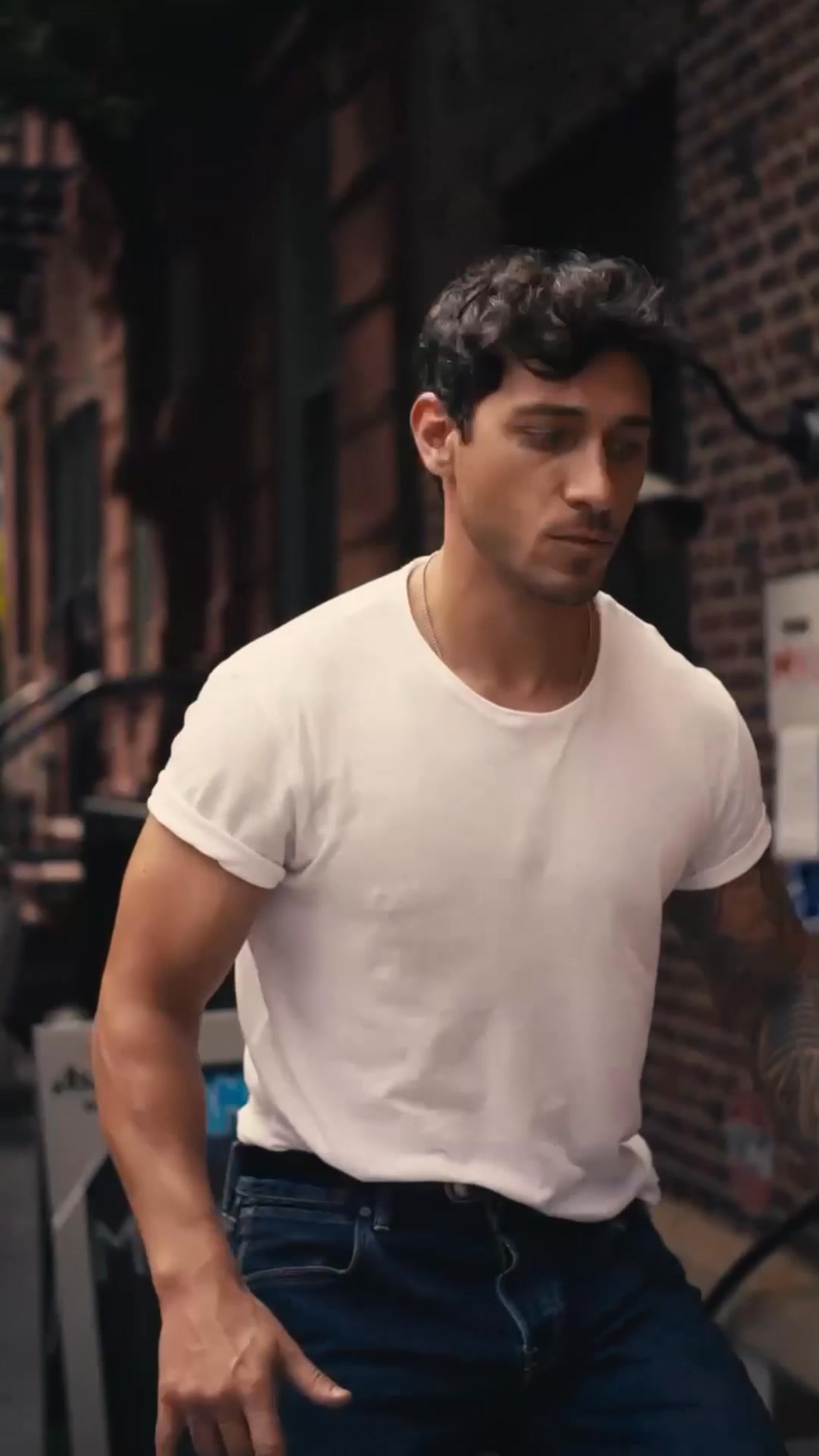 A man with dark, curly hair and a white t-shirt is shown from the chest up, looking down and to the side. He is wearing blue jeans and a thin necklace. The background is a blurred urban street scene with brick buildings. Text overlaying the image reads: "Rob Raco wears the Autumn ’25 menswear collection – shop it now."