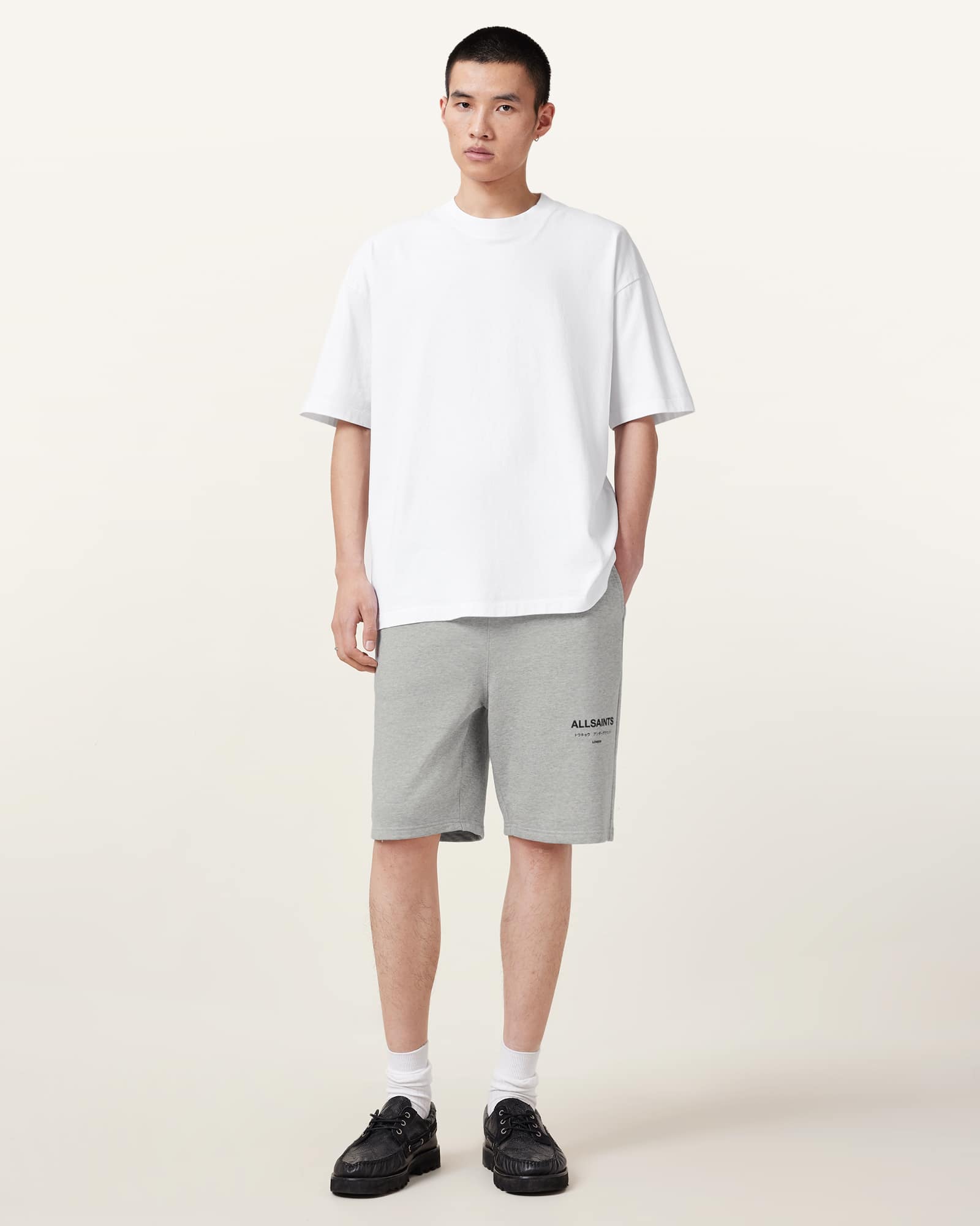 A male model stands centered against a plain white background, wearing a white t-shirt and grey shorts with the AllSaints logo. He is looking directly at the camera with his hands in his pockets. The text "Shop the AllSaints Sale now, plus free returns." is overlaid on the image in black sans-serif font.