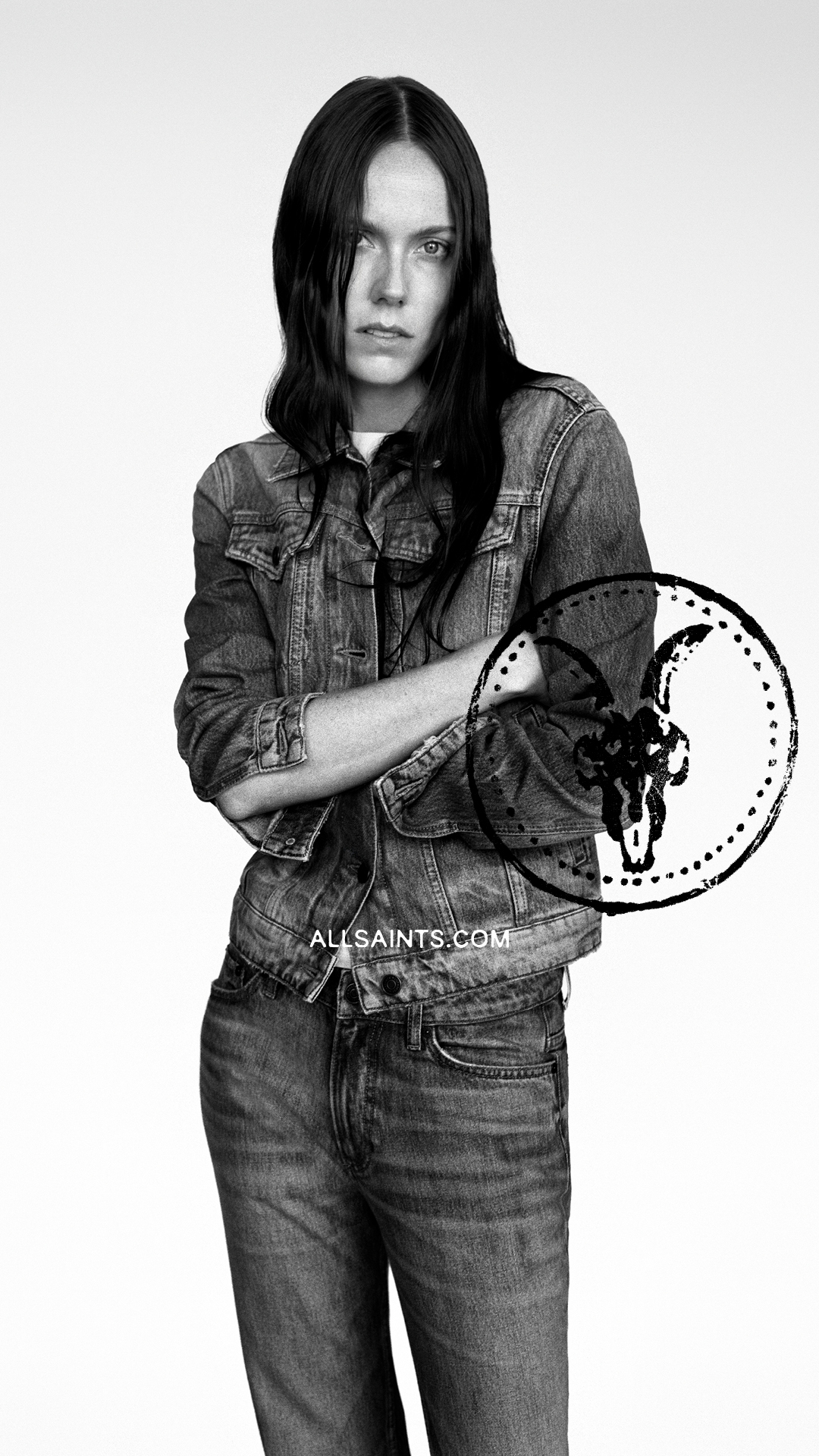 A black and white, medium shot of a young woman with long, dark hair, wearing a denim jacket and jeans. She is standing with her arms crossed and looking directly at the camera with a serious expression. The AllSaints logo is visible on the bottom of the image. The background is plain white.