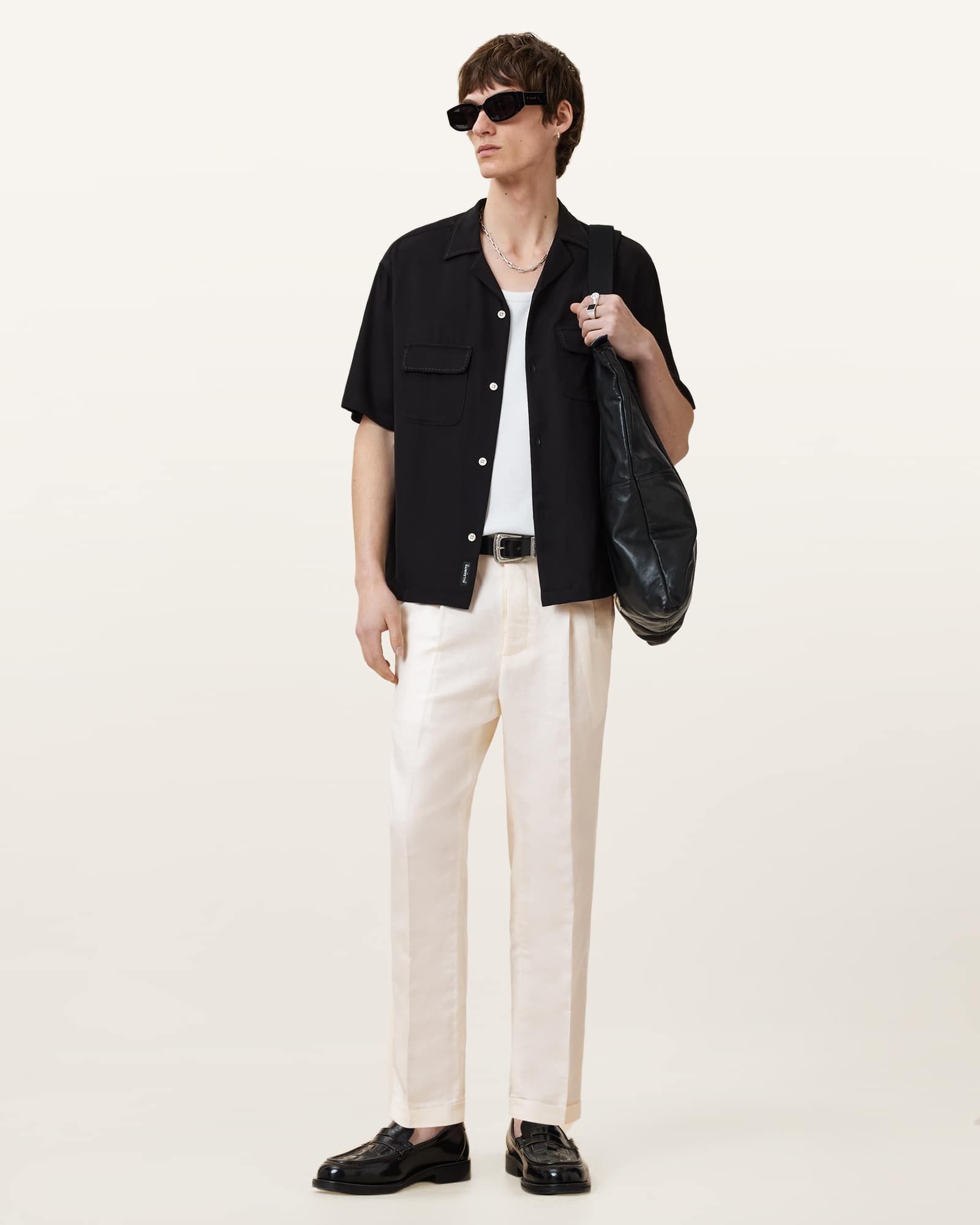 A full-body shot of a male model wearing a black short-sleeved button-up shirt over a white t-shirt, cream-colored pleated trousers, and black loafers. He is accessorized with sunglasses, a silver chain necklace, a silver ring, and a black leather shoulder bag. The background is a plain, light-colored studio setting. The text overlay advertises a sale with free delivery over £100.