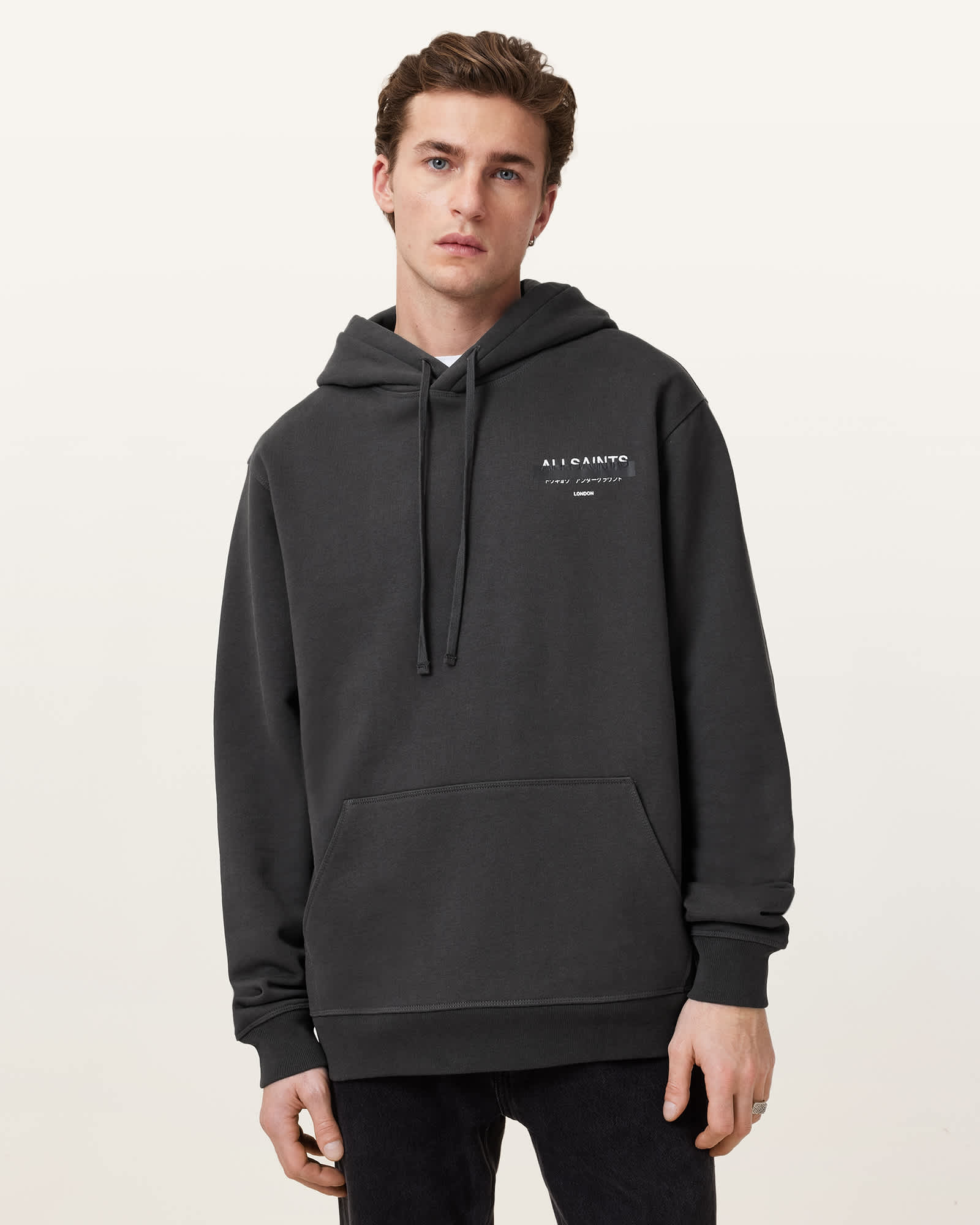A man stands facing the camera wearing a dark grey hooded sweatshirt with a small white logo and text on the left chest. The background is a plain light grey. The text on the sweatshirt reads "ALLSAINTS" with smaller text below it. The ad copy is overlaid on the image, stating "New-Season ALLSAINTS has landed. Shop now & get free delivery over £100." The man is looking directly at the viewer with a neutral expression.