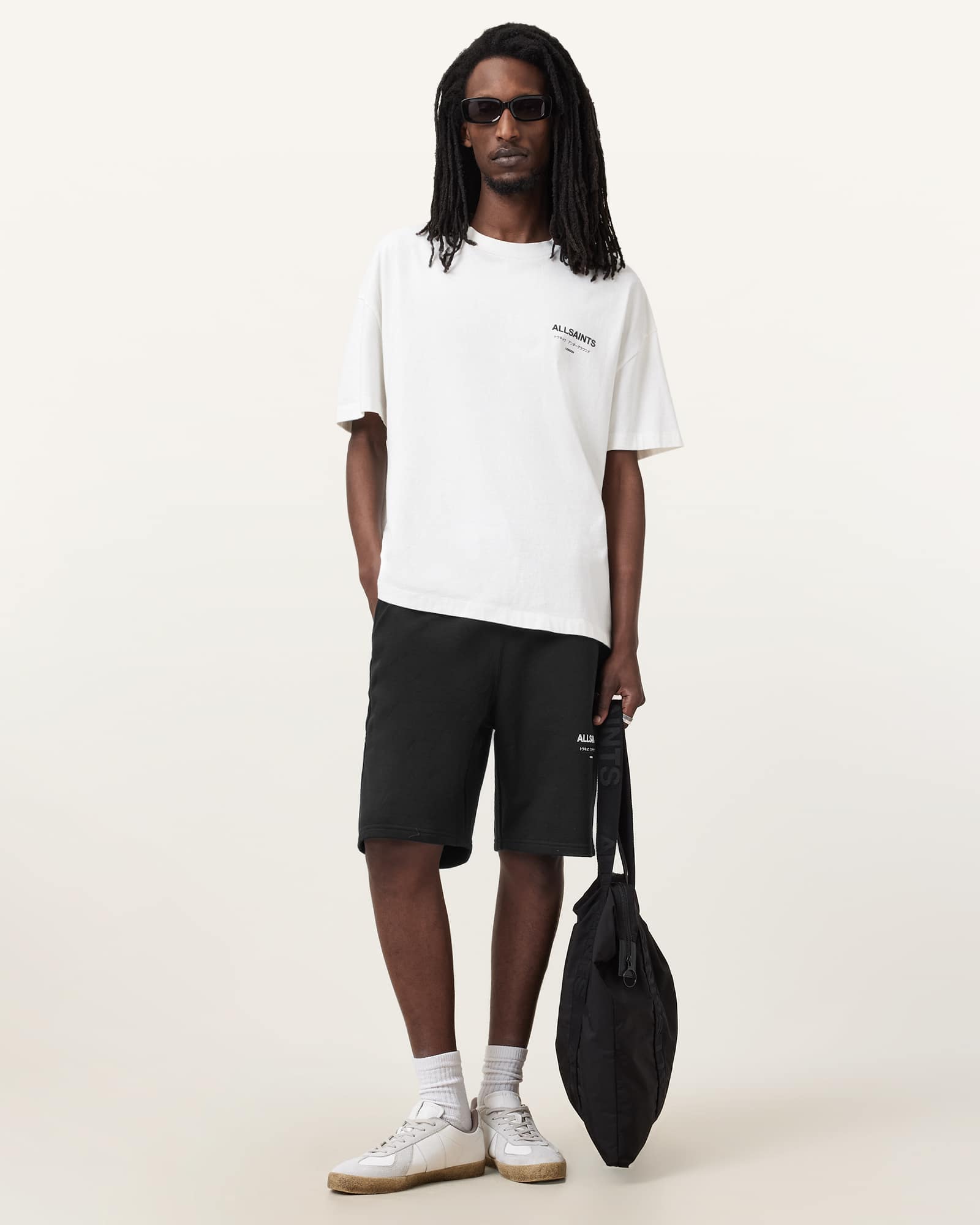 A male model stands against a plain background, wearing a white t-shirt with a small black logo and black shorts. He is also wearing sunglasses, white socks, and light-colored sneakers, and is holding a black tote bag. The text "It's here – shop new-season AllSaints now and enjoy free delivery over £100." is overlaid on the image. The overall presentation is clean and highlights the new season's collection with a delivery offer.
