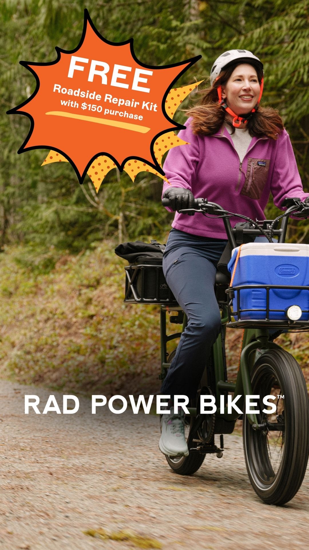 A woman wearing a helmet and a purple fleece jacket rides an e-bike on a paved path through a wooded area. She is smiling and looking to the side. A graphic overlay in the top left corner reads "FREE Roadside Repair Kit with $150 purchase". The text "RAD POWER BIKES" is displayed in large white letters across the bottom of the image.