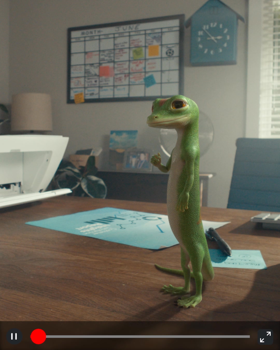 A CGI gecko stands on a wooden desk in an office setting. Behind him, a calendar for June is visible on the wall, along with a blue clock. A blue document with a diagram is spread out on the desk in front of the gecko. The gecko is looking towards the viewer and has its right hand raised with a thumb up. The text "GEICO makes home insurance easy. So you can fast forward through all the stuff you don't need." is overlaid on the image.
