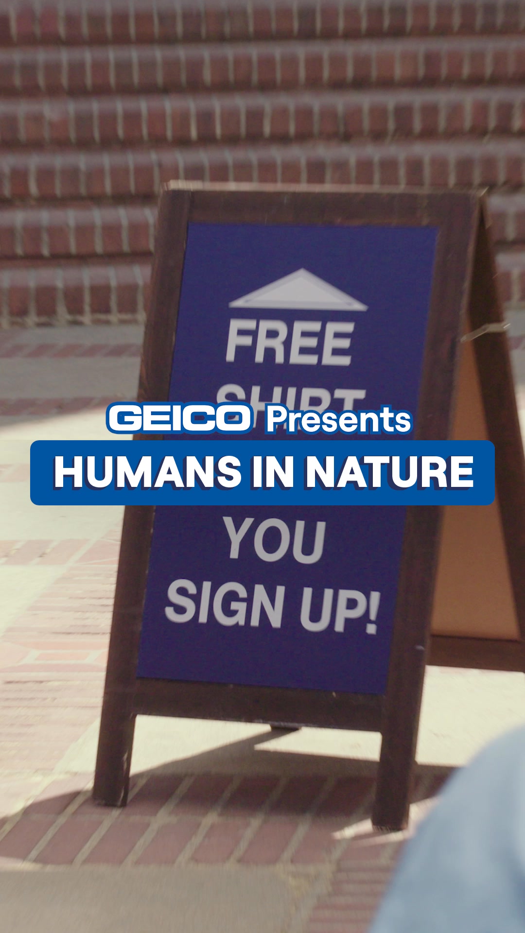 A wooden A-frame sign stands on a brick walkway. The sign has a dark blue background with white text. The top of the sign reads "FREE SHIRT" with a white house icon above it. Below that, "GEICO Presents" is written in smaller text. The main message on the sign is "HUMANS IN NATURE YOU SIGN UP!". The creative uses a contrast hook, implying that the money saved with GEICO is more valuable than a free shirt.