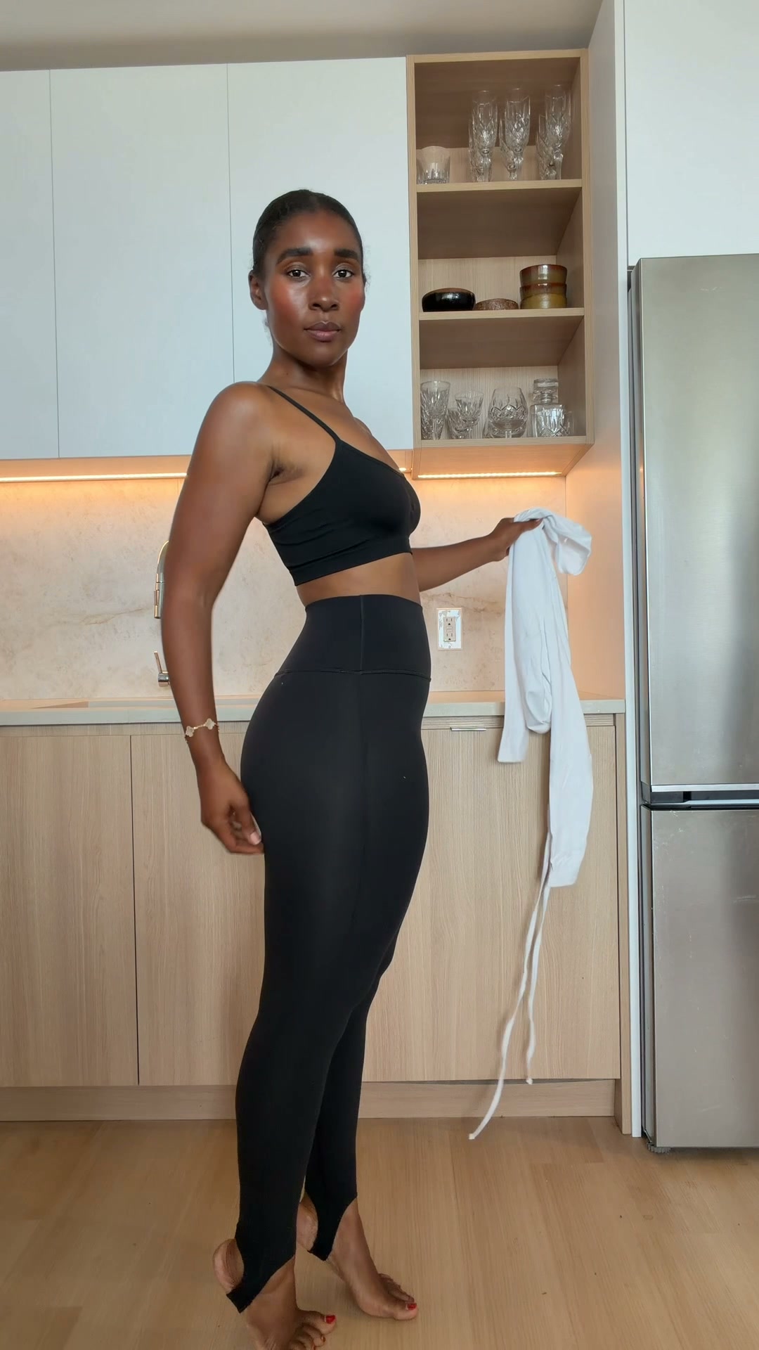 A woman wearing black athleisure clothing, including stirrup leggings and a sports bra, stands in a modern kitchen. She is holding a white garment in her right hand. The text "From your first plan to your last. Made to go with you everywhere." is overlaid on the image. The overall aesthetic suggests comfort and versatility for an active lifestyle.