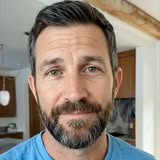 A close-up selfie video of a man with short brown hair and a beard, wearing a blue t-shirt. He is looking directly at the camera with a slight smile. The background appears to be a home interior with wooden beams visible on the ceiling and cabinets in the distance. The on-screen text reads: "I'm about to mansplain perimenopause." The creator is speaking directly to the viewer, adopting a conversational and slightly humorous tone to introduce a topic that might be considered outside of typical male discourse.