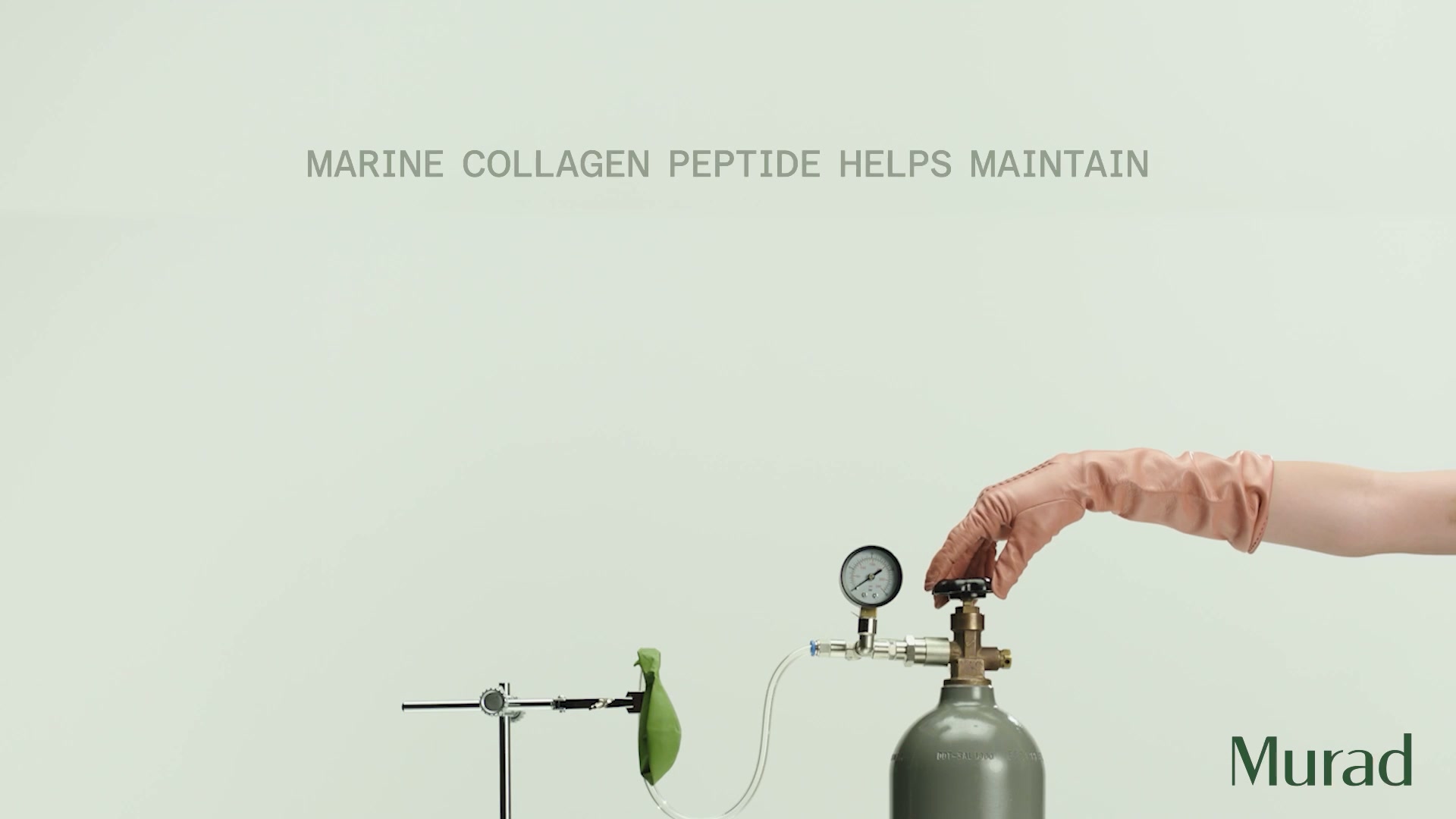 The creative features a light grey background with text at the top that reads "MARINE COLLAGEN PEPTIDE HELPS MAINTAIN". In the lower right corner, the Murad logo is visible. The main visual shows a hand wearing a light pink glove, turning a valve on a grey gas tank. A clear tube connects the tank to a small, green, deflated balloon which is attached to a stand on the left side of the image. A pressure gauge is attached to the valve assembly, showing a reading.