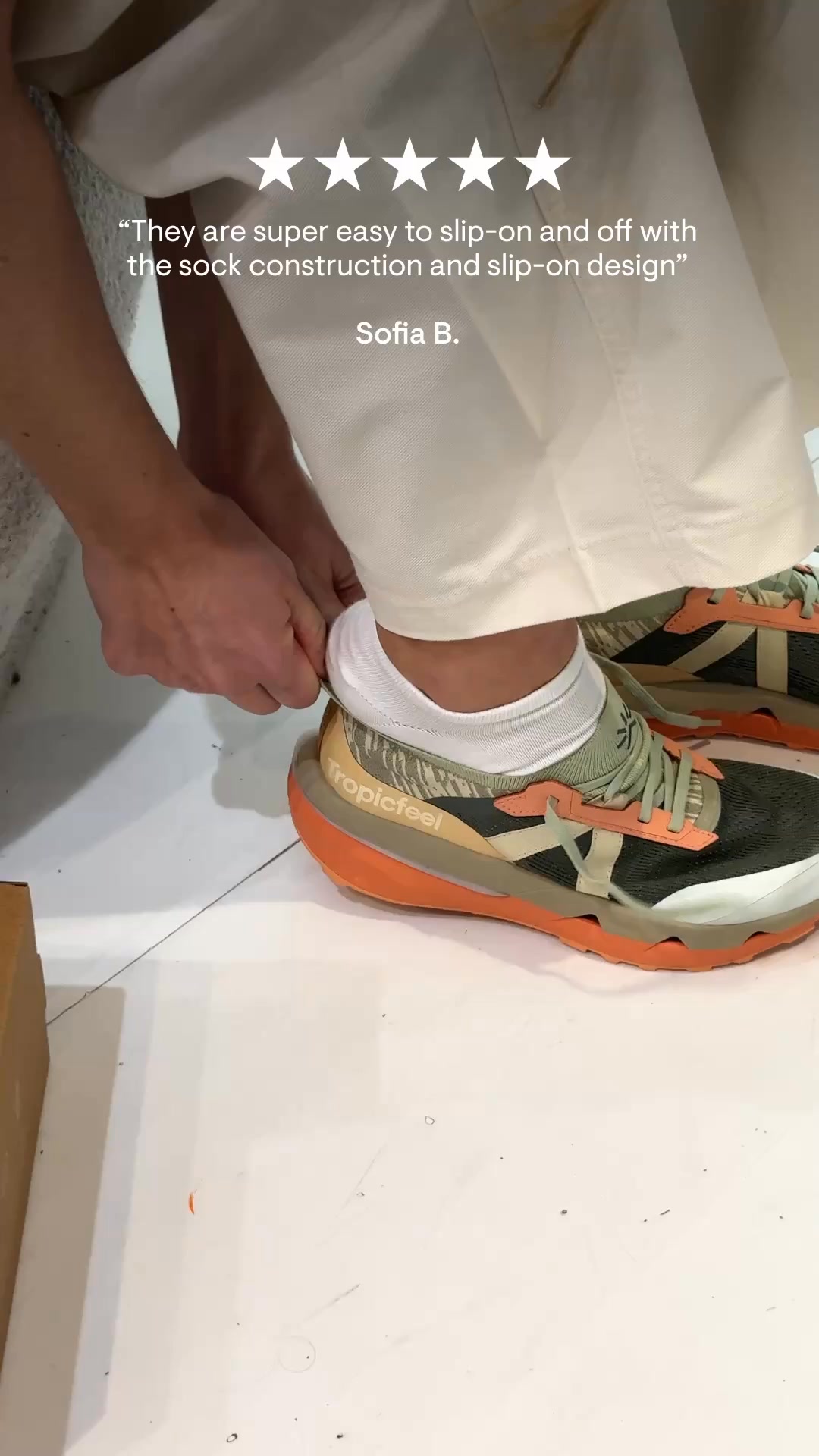 A close-up shot shows a person putting on a shoe. The person is wearing white socks and off-white pants. The shoe is a dark green and orange sneaker with the brand name "Tropicfeel" visible on the side. Five white stars are displayed above a quote attributed to "Sofia B.", which reads: “They are super easy to slip-on and off with the sock construction and slip-on design”.
