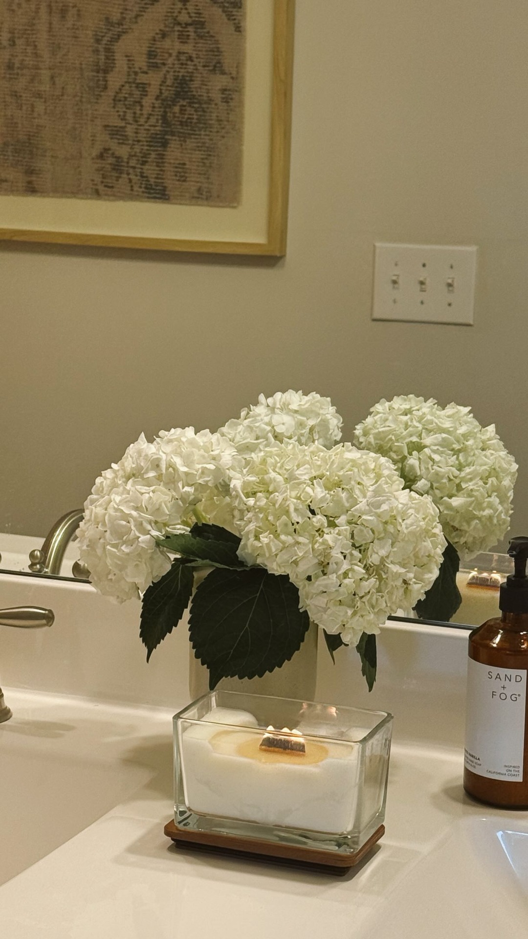 A clean and minimalist bathroom vanity is shown, featuring a white sink, a modern faucet, a vase of white hydrangeas, and a lit square candle in a glass container. The text "anyway, we’re bidet people now" is overlaid on the image. The overall aesthetic suggests a serene and well-appointed personal space.