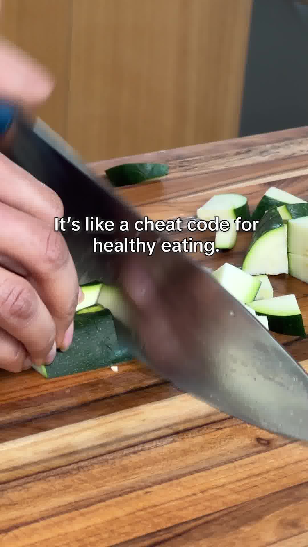 A close-up shot shows a person's hands chopping zucchini on a wooden cutting board. Sliced zucchini pieces are scattered around the board. White text overlays the image, reading "It's like a cheat code for healthy eating." The overall scene suggests a quick and easy way to prepare healthy food.
