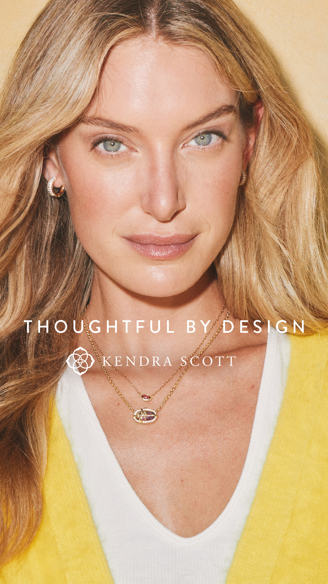 A close-up shot of a blonde woman wearing a yellow cardigan over a white top, with delicate gold necklaces. The text "THOUGHTFUL BY DESIGN" is overlaid in white, followed by the Kendra Scott logo and name. The image focuses on the jewelry and the model's serene expression, highlighting the brand's aesthetic and the concept of thoughtful design in their products.