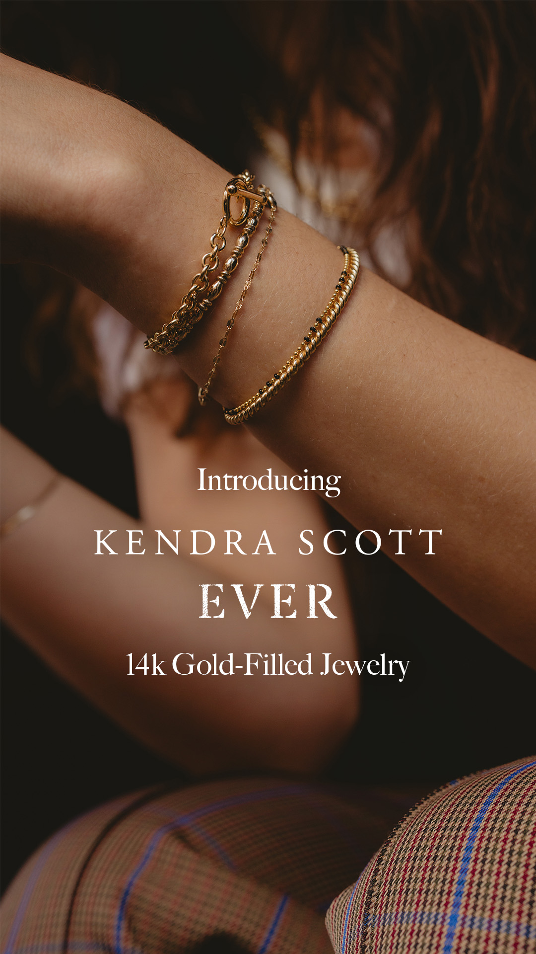 A close-up shot focuses on a person's arm adorned with multiple gold bracelets. The background is softly blurred, with hints of dark hair and plaid fabric. White text overlays the image, announcing "Introducing KENDRA SCOTT EVER" and "14k Gold-Filled Jewelry," highlighting the brand and the product line.