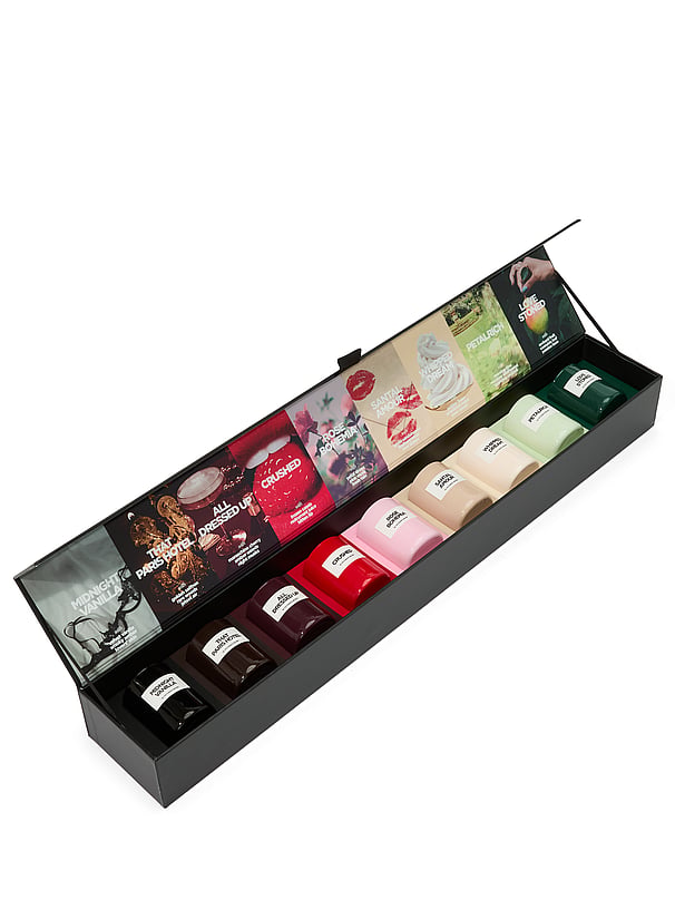 A black gift box is open, revealing a collection of small, colorful perfume bottles arranged in two rows. The lid of the box is propped open, displaying a collage of images and text related to the scents. The bottles are labeled with names such as "Midnight Vanilla," "All Dressed Up," and "Crushed." The overall presentation suggests a curated selection of fragrances, possibly for gifting or personal exploration.