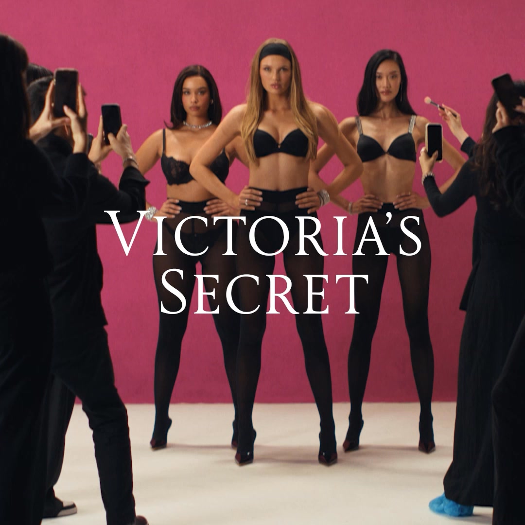 Three models are posed in black lingerie and stockings against a bright pink backdrop. The Victoria's Secret logo is prominently displayed in white text across the center of the image. The models are being photographed by several people holding up smartphones, and one person is applying makeup to the model on the right. The text overlay states, "Our bras prove it. Discover the most iconic models ever created."