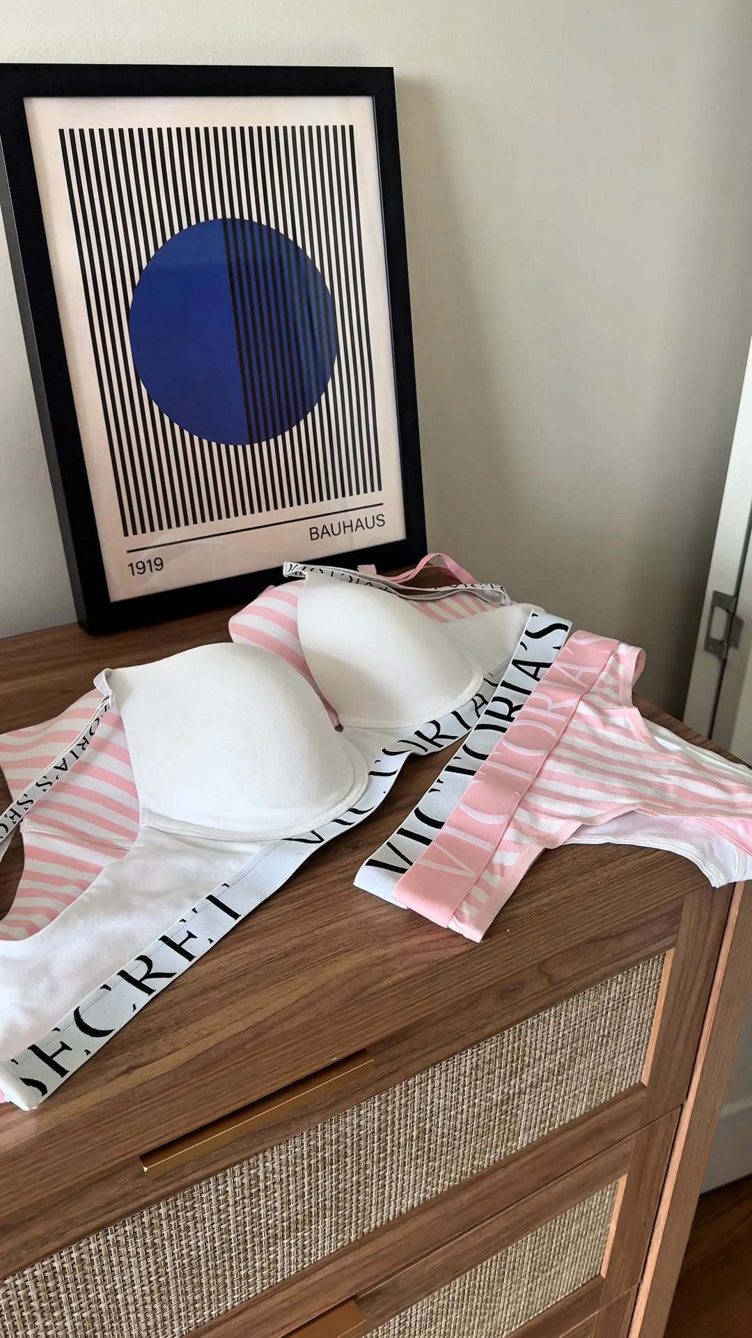A white Victoria's Secret bra and matching pink and white striped underwear are laid out on a wooden dresser. The bra has the brand's logo printed in black on its straps, and the underwear also features the logo in pink. Behind the lingerie, a framed abstract art print with black stripes and a blue circle is leaning against the wall. The text overlay highlights the bra's improved design for invisibility under clothing.