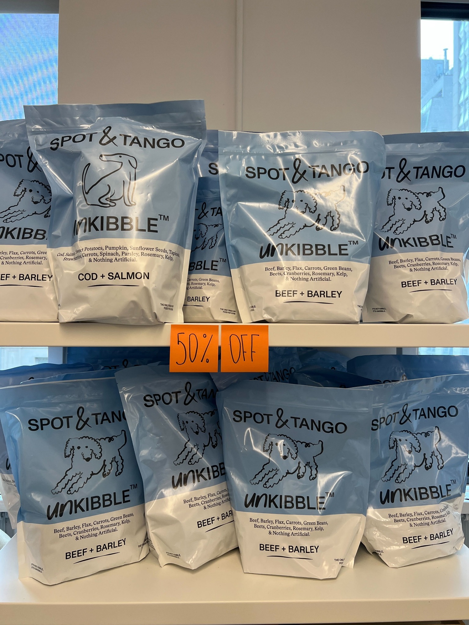 The image displays multiple bags of Spot & Tango dog food, labeled 'UnKibble'. The bags are light blue with white text and illustrations of dogs. Some bags show different flavors like 'Cod + Salmon' and 'Beef + Barley'. An orange sign with '50% OFF' written in black marker is prominently displayed in the center of the image, indicating a promotion. The overall presentation is a product-focused shot with a clear offer highlighted.