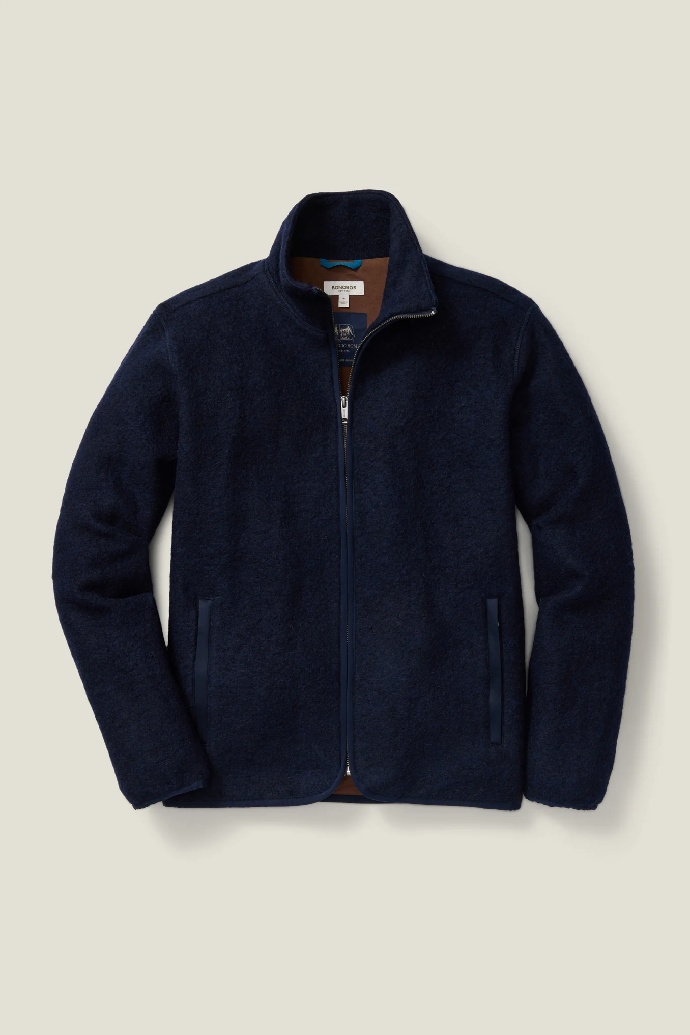 A dark navy fleece jacket is shown laid flat against a light beige background. The jacket is zipped up, revealing a brown interior lining and a Bonobos brand tag. The image is a clean, studio shot of the product. Text overlays the image, stating "Meet the building blocks for creating a great-fitting wardrobe that feels like it was designed just for you. Complimentary shipping & returns in the U.S."