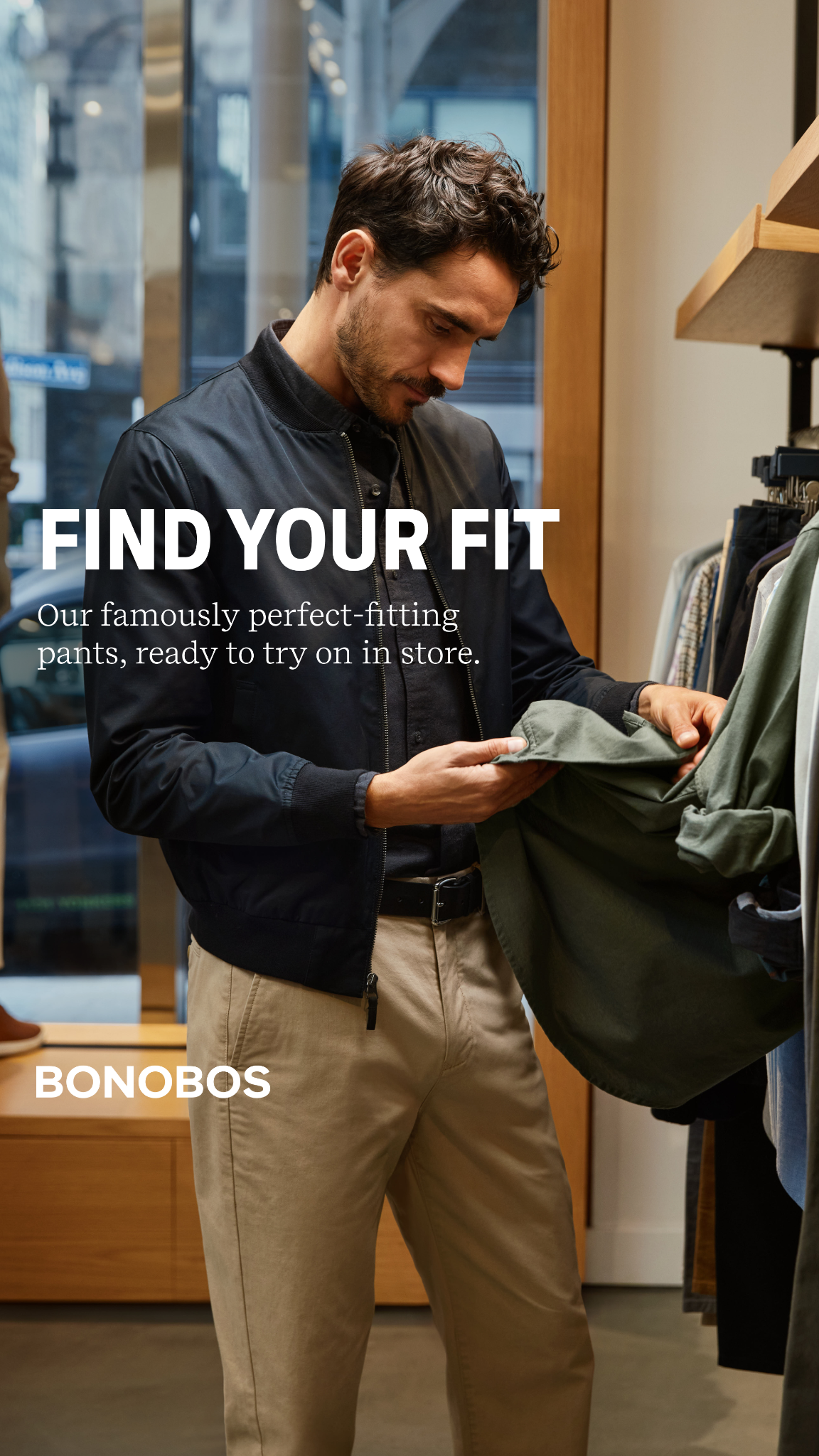 A man in a dark bomber jacket and tan pants is in a store, looking down at a green shirt he is holding. White text on the left side of the image reads "FIND YOUR FIT" and below that, "Our famously perfect-fitting pants, ready to try on in store." The Bonobos logo is visible in white text at the bottom left corner of the image.