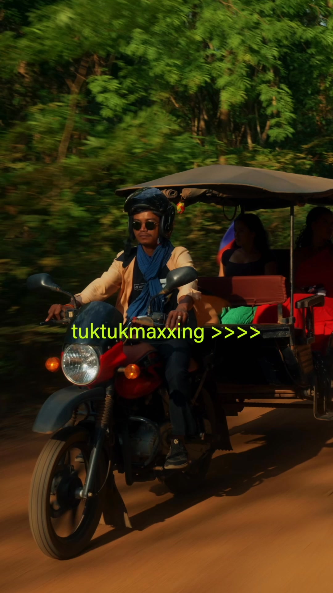 A man wearing sunglasses and a blue scarf rides a tuk-tuk down a dirt road, with two passengers behind him. The background is filled with lush green trees. The text "tuktukmaxxing >>>" is overlaid on the image in bright green.
