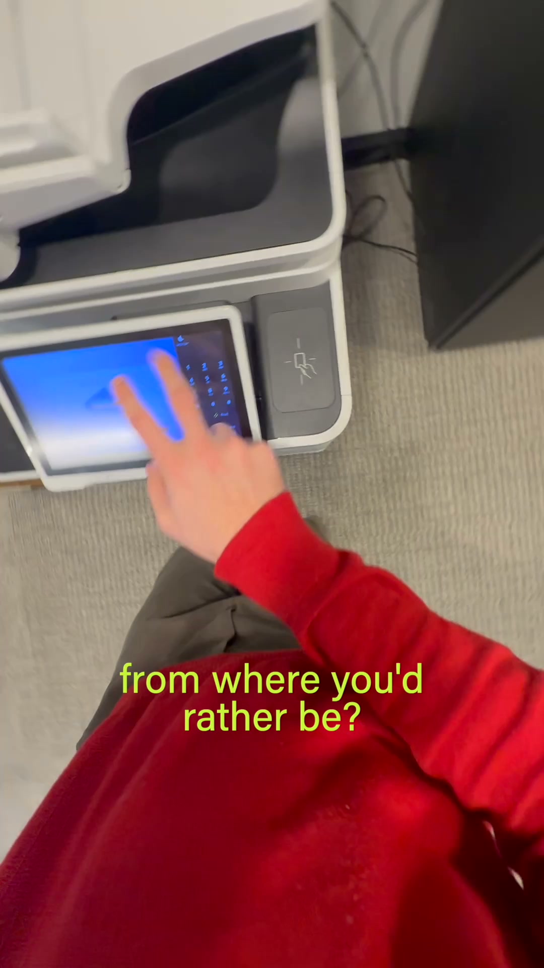 A person wearing a red sweatshirt and grey pants is shown from the chest down, interacting with a touchscreen interface on a printer. The screen displays a keypad and a blue background. Bright yellow text overlaid on the image asks, "from where you'd rather be?" The scene suggests a moment of contemplation or a choice between the current activity and a more desirable location.
