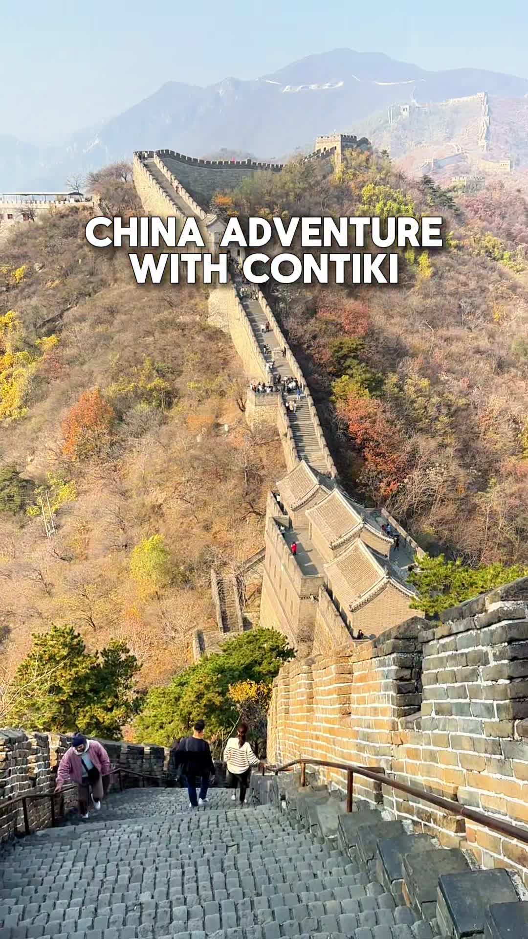 A wide shot of the Great Wall of China, with a steep stone staircase in the foreground and a long, winding section of the wall stretching into the distance. Several tourists are visible walking on the wall. Large white text overlaid on the image reads "CHINA ADVENTURE WITH CONTIKI". The surrounding hills are covered in dry brush and scattered trees with autumn foliage.