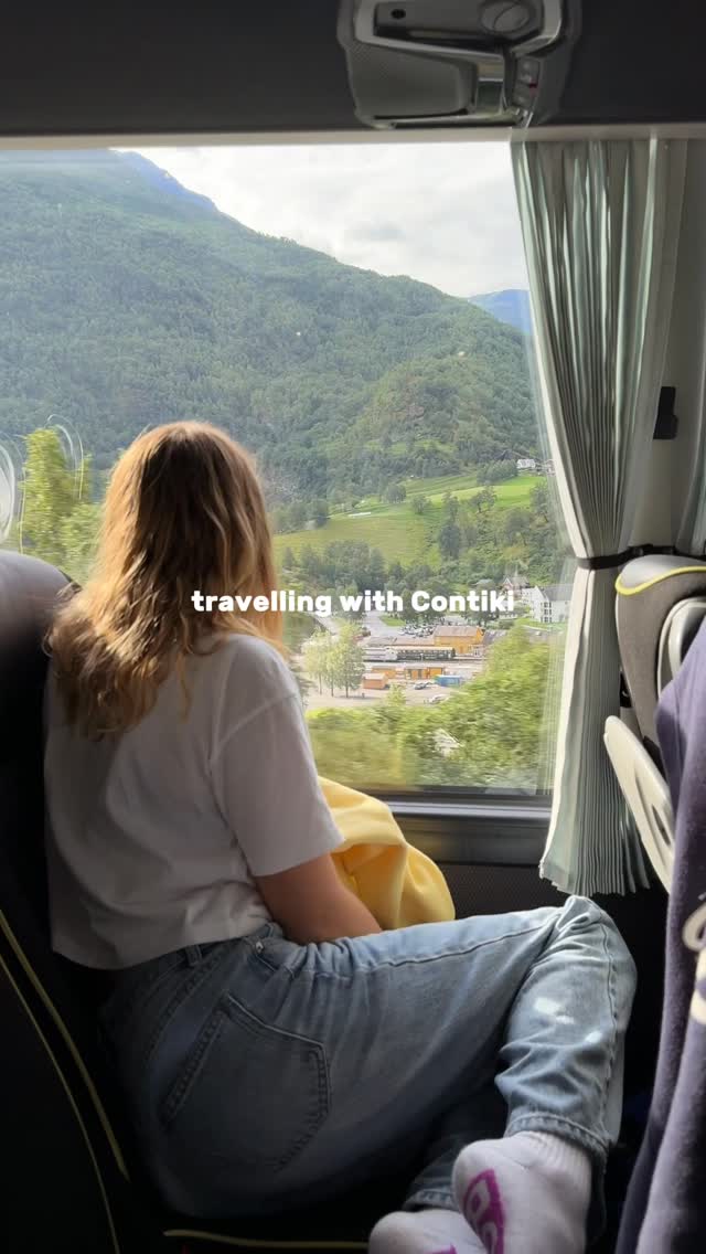A young woman with blonde hair sits on a bus, looking out the window at a scenic mountain landscape. The text "travelling with Contiki" is overlaid on the image. She is wearing a white t-shirt and blue jeans, with a yellow cloth draped over her lap. The overall impression is one of relaxed travel and enjoying the view.