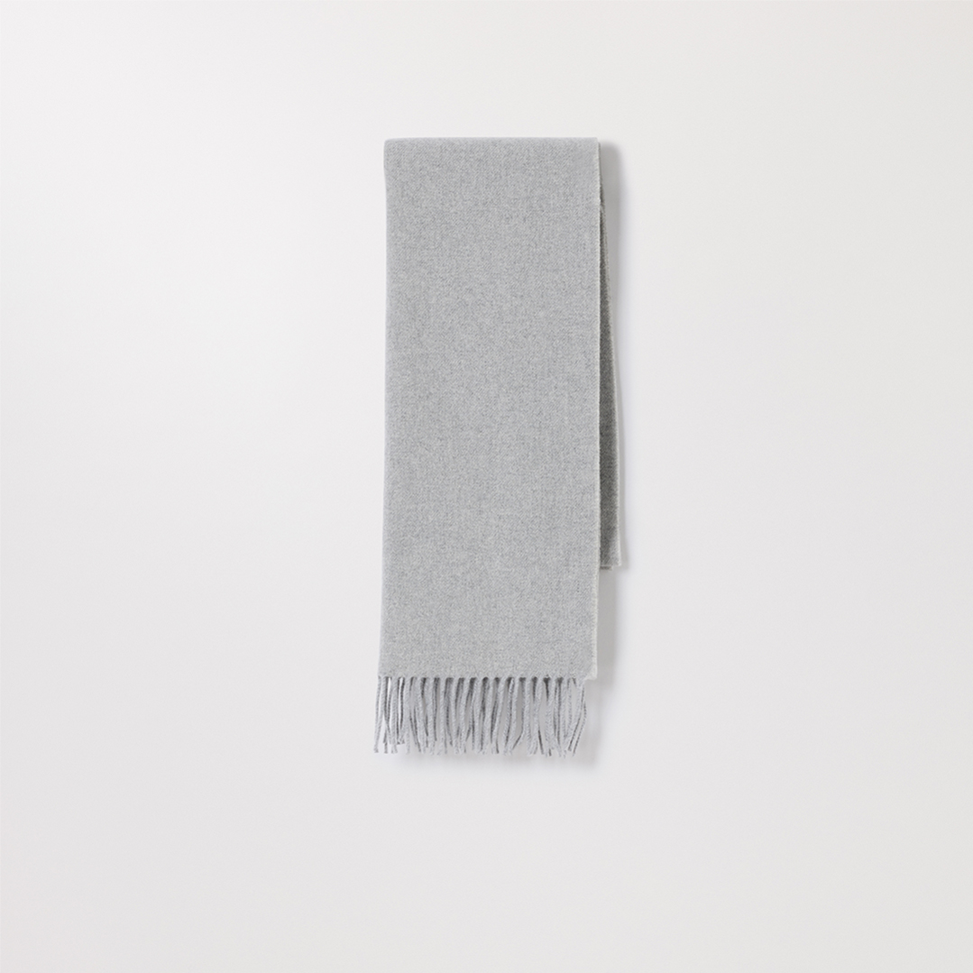 A light grey scarf with fringe on the bottom is displayed against a plain white background. The scarf is folded in half lengthwise and hangs vertically. There is no text on the image itself, but the copy provides context about a permanent collection for women. The visual focuses solely on the product's presentation.