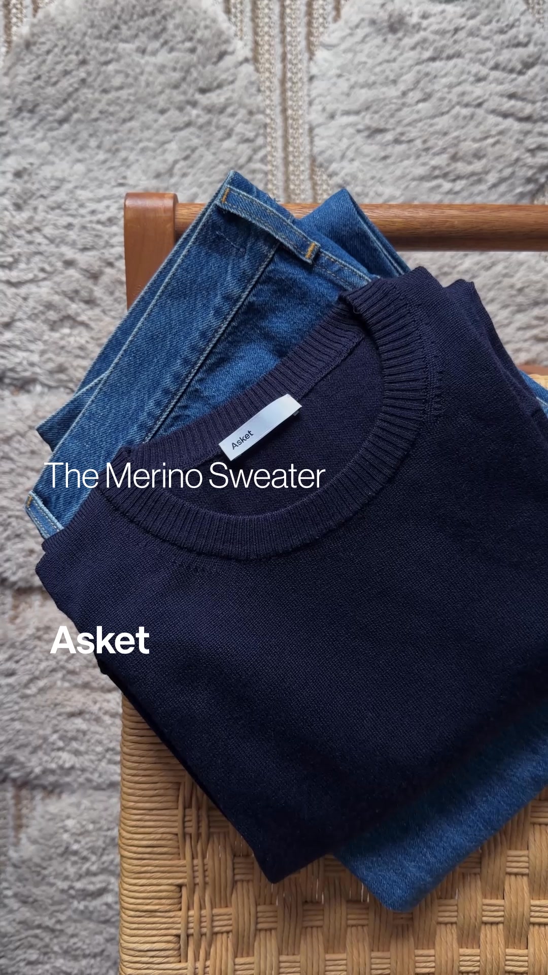 A dark navy merino wool sweater is folded on top of a pair of blue denim jeans, which are resting on a woven chair. The scene is set against a textured rug. White text overlays the image, with the brand name 'ASKET' at the bottom left and 'The Merino Sweater' positioned above the sweater's neckline. The sweater has a small white tag with the ASKET logo.