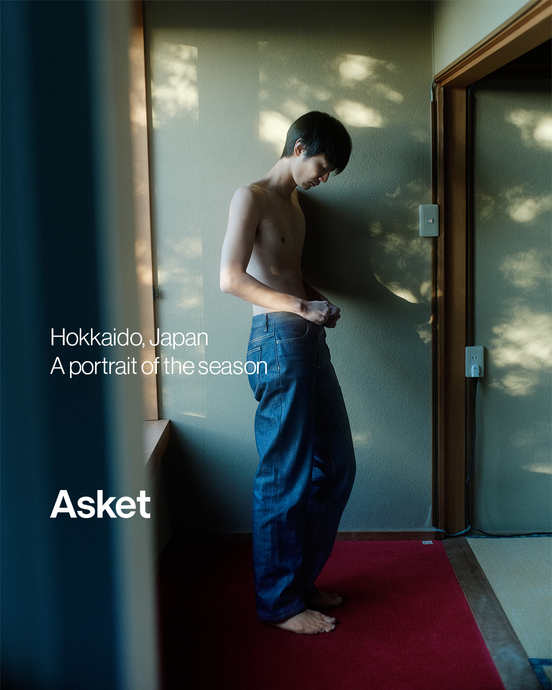 A young man, shirtless and wearing loose-fitting dark denim jeans, stands in a room with soft, dappled sunlight filtering through a window. He is looking down and adjusting his jeans. White text on the left side of the image reads "Hokkaido, Japan" and "A portrait of the season", followed by the brand name "Asket" in larger white font. The overall mood is introspective and serene, capturing a quiet moment in a specific location.
