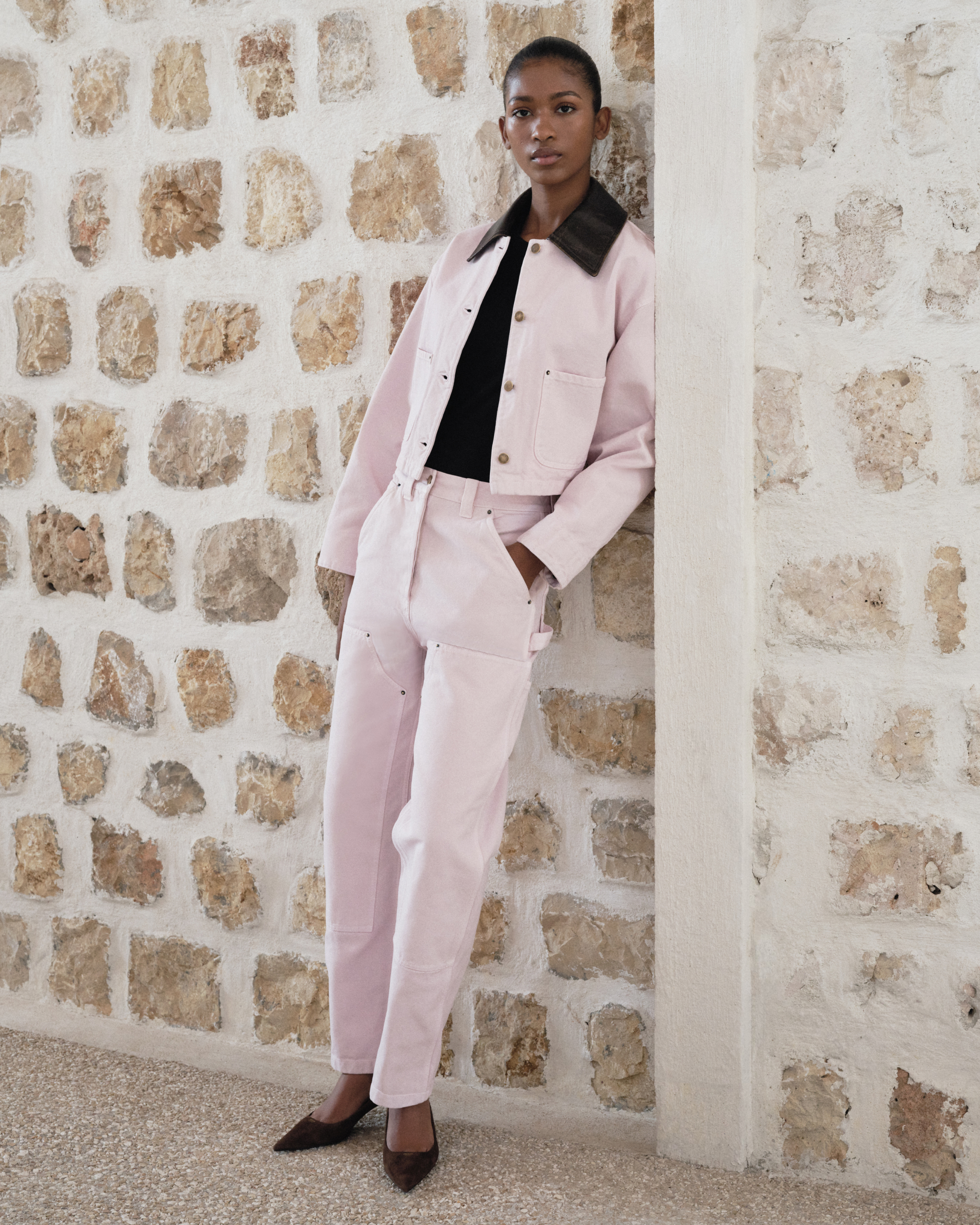 A young Black woman stands leaning against a textured stone wall, wearing a light pink denim jacket and matching pants over a black top. She is looking towards the camera with a neutral expression. The text "This just in: shop new styles from Prada now on FARFETCH" is overlaid on the image. The overall aesthetic is minimalist and chic, highlighting the fashion ensemble.