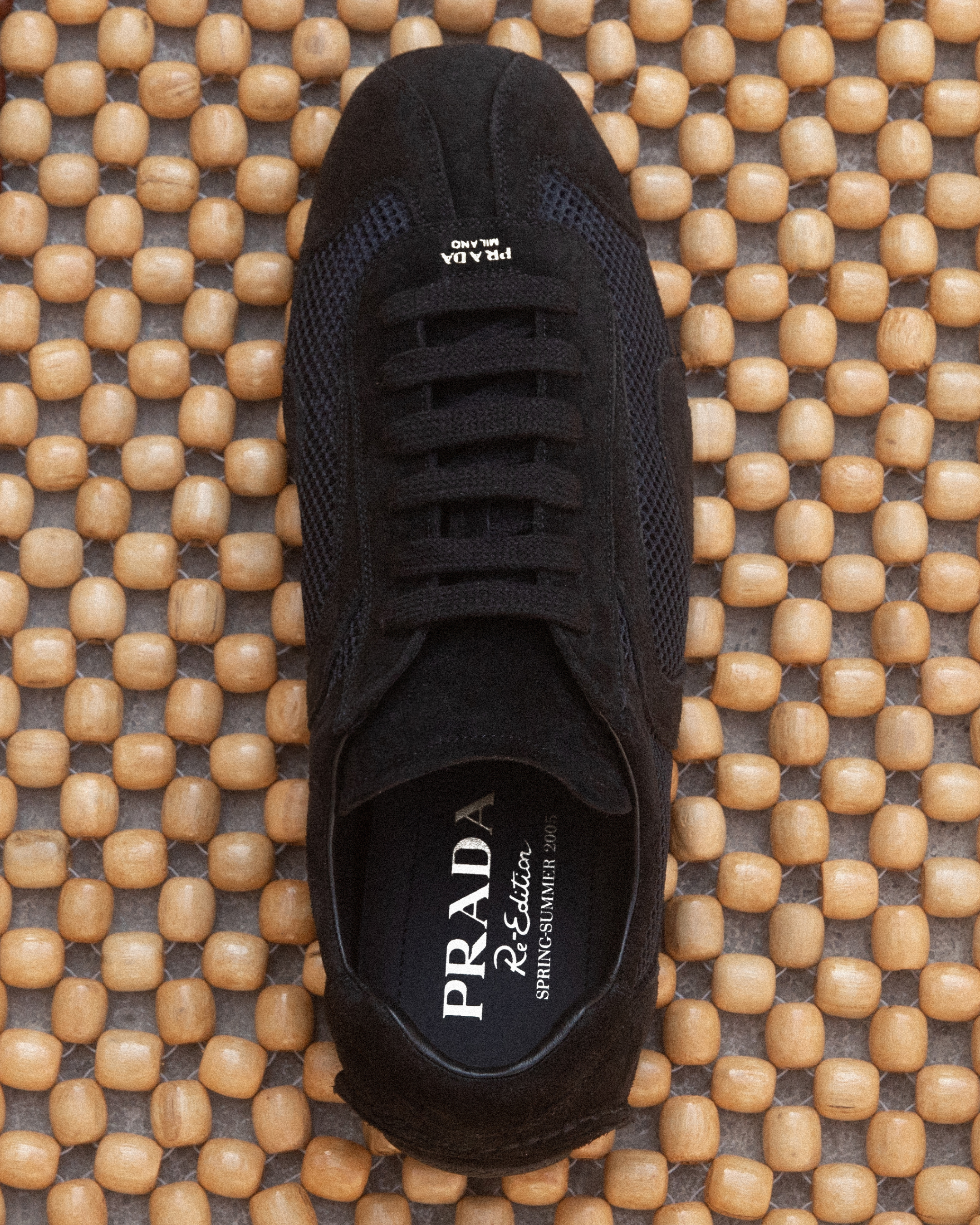 A top-down view of a black Prada sneaker is displayed against a background of light brown wooden beads. The shoe features a mesh and suede construction with black laces. The Prada logo and 'Re-Edition SPRING-SUMMER 2005' are visible on the insole. The text 'New-season Prada? Farfetch it.' is overlaid on the image.