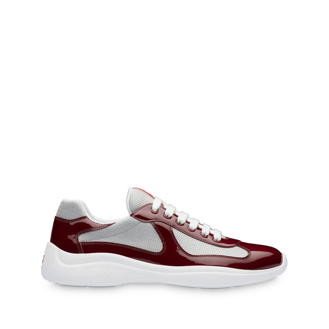 A studio shot of a single Prada sneaker is presented against a white background. The sneaker is a low-top design with a burgundy patent leather and grey mesh upper, white laces, and a thick white sole. There is no text overlay on the image itself, but the ad copy mentions exploring Prada Shoes on FARFETCH.