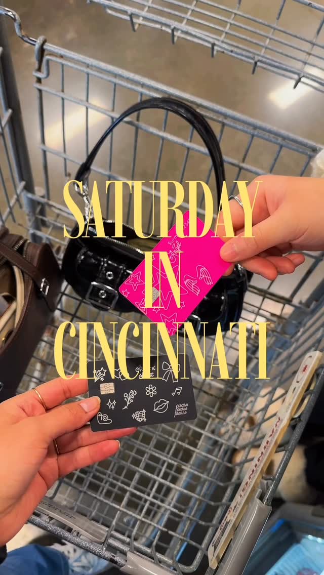 The creative shows a shopping cart with two hands holding up credit cards. The text "SATURDAY IN CINCINNATI" is overlaid in large yellow font. One credit card is bright pink with white angel wing and star graphics. The other credit card is black with white line drawings of various icons, including a snail, a flower, lips, and a globe. The overall scene suggests a shopping trip or a day out.