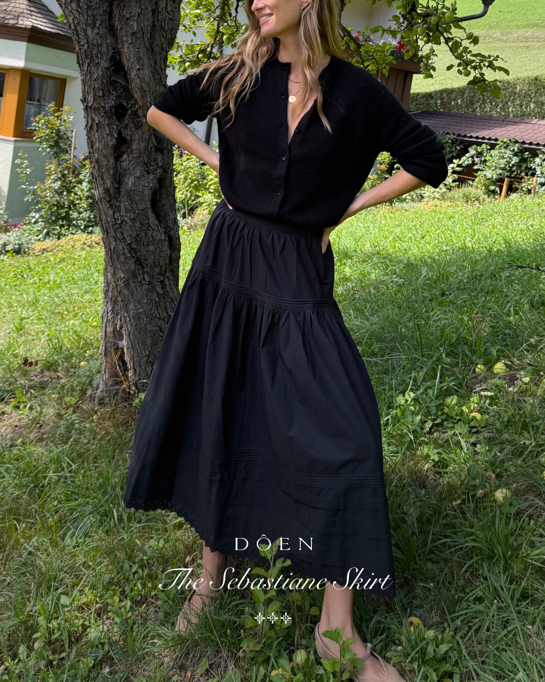 A woman stands in a grassy outdoor setting, leaning against a tree. She is wearing a black cardigan and a long, flowing black skirt. The text "DÔEN The Sebastiane Skirt" is overlaid on the image. The copy explains that the skirt is inspired by an Edwardian-era trousseau and made of organic cotton poplin.