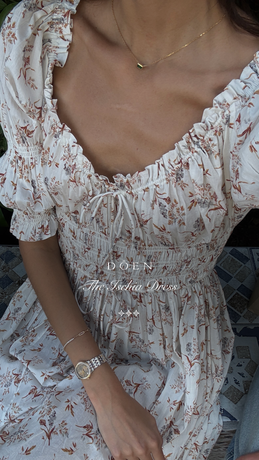 A close-up shot of a woman wearing a floral print dress. The dress has a ruffled neckline and smocked bodice. Text overlaid on the image reads "DOEN The Ischia Dress" with decorative symbols. The woman is wearing a delicate necklace and a watch on her left wrist. The overall aesthetic is romantic and feminine, suggesting the dress is suitable for various styling options throughout the year.