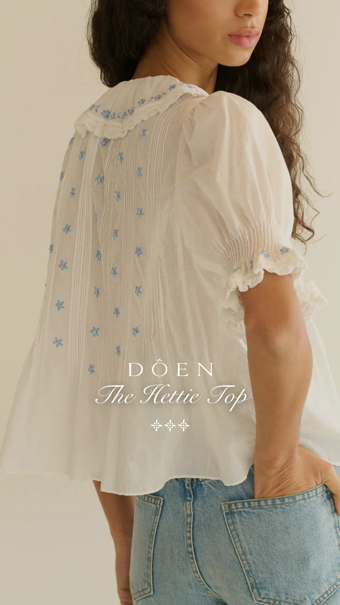A woman is shown from the back, wearing a white, embroidered blouse and light-wash denim jeans. The blouse features delicate blue floral embroidery on the collar and scattered across the fabric, with smocked details on the sleeves. The brand name "DÔEN" and the product name "The Hettie Top" are displayed in white text over the blouse. The copy highlights the blouse's versatility and material.