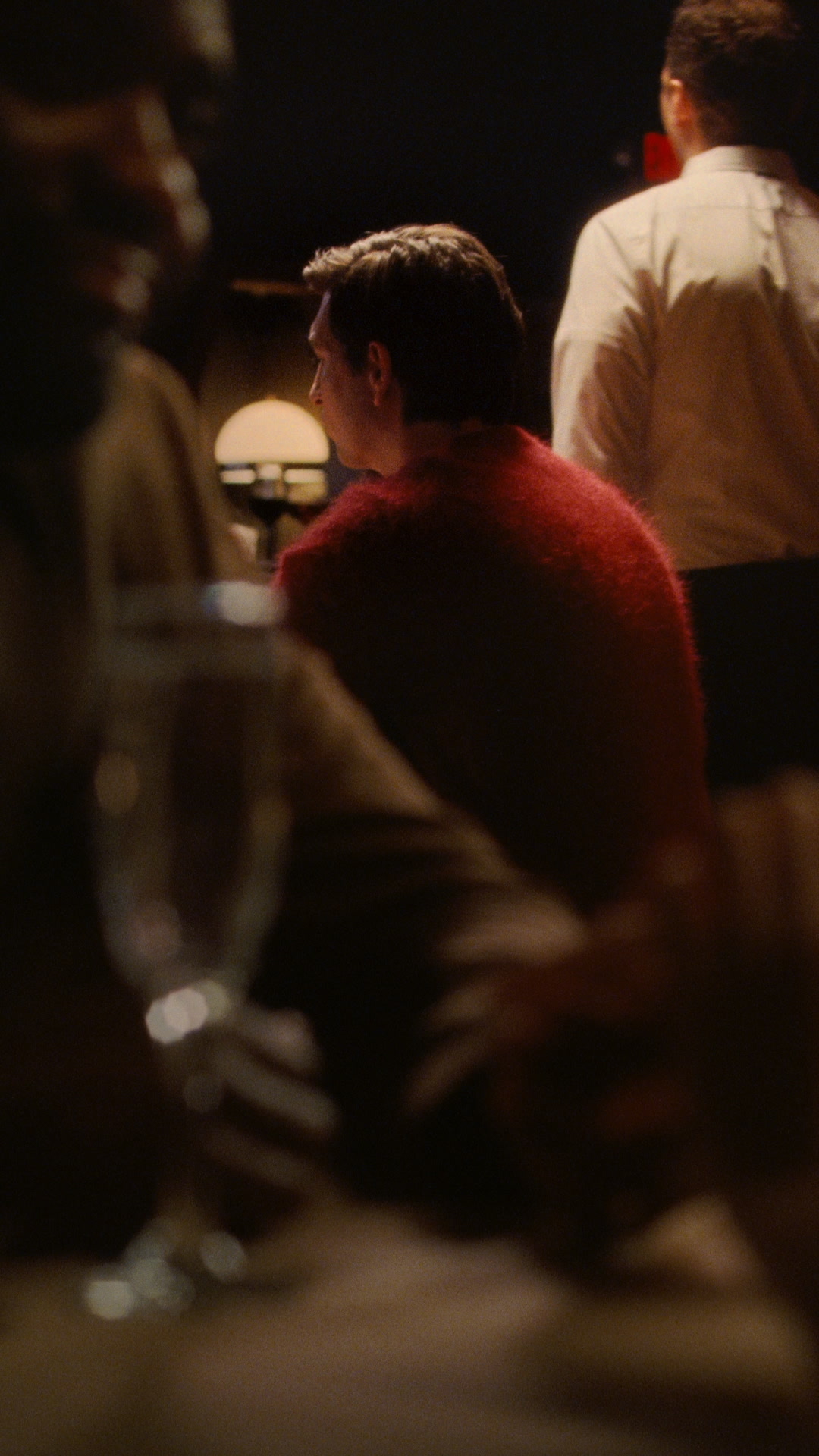 The image shows a dimly lit, intimate gathering with several people in the foreground and background. A man in a red fuzzy sweater is the central focus, seen from the side as he looks away. Another person in a white shirt is visible behind him. The text overlay reads "Your time belongs to you. Set the pace with the Runwell, assembled in Detroit." The overall mood is sophisticated and relaxed.