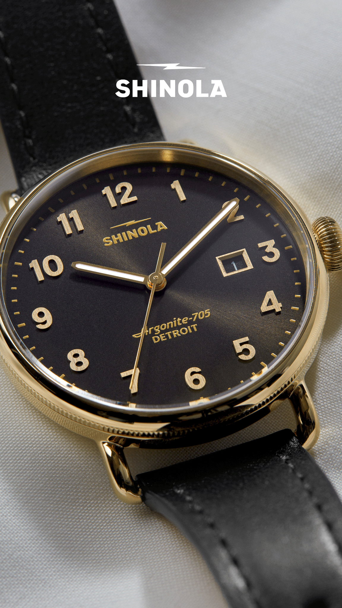 A close-up, detailed shot of a Shinola watch with a black dial and gold casing. The watch face displays gold hour markers and hands, with the brand name 'SHINOLA' prominently featured. Text overlays the image, describing the watch as a 'quietly confident, dressier Canfield' with specific dimensions and color details. The background is a soft, textured white fabric, drawing full attention to the timepiece.