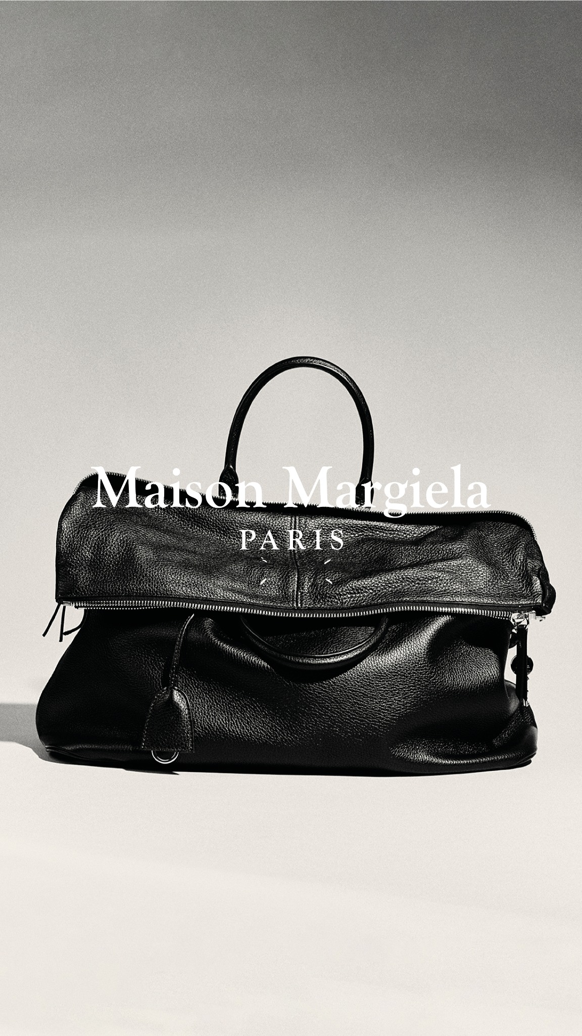 A black leather Maison Margiela handbag is displayed against a neutral background. The brand name "Maison Margiela" is overlaid in white text at the top of the image, with "PARIS" centered below it. The text explains that the selection of bags features established and new designs, with each piece reflecting the brand's approach to materials and construction.