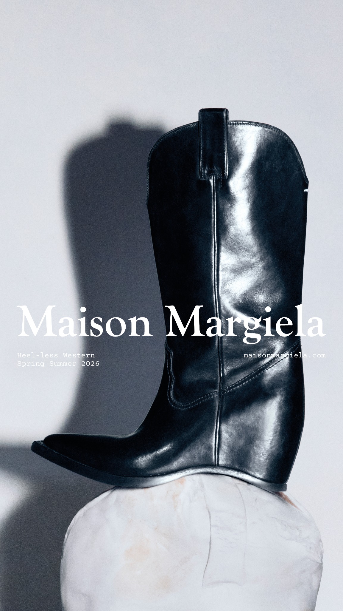 A high-fashion, black leather cowboy boot is displayed on a white, abstract pedestal against a grey background. The Maison Margiela logo is prominently displayed in white text on the left side of the image, with the collection name "Heel-less Western Spring Summer 2026" below it. The website address "maisonmargiela.com" is also visible on the right side. The creative focuses on the unique design of the boot, highlighting its heel-less construction.