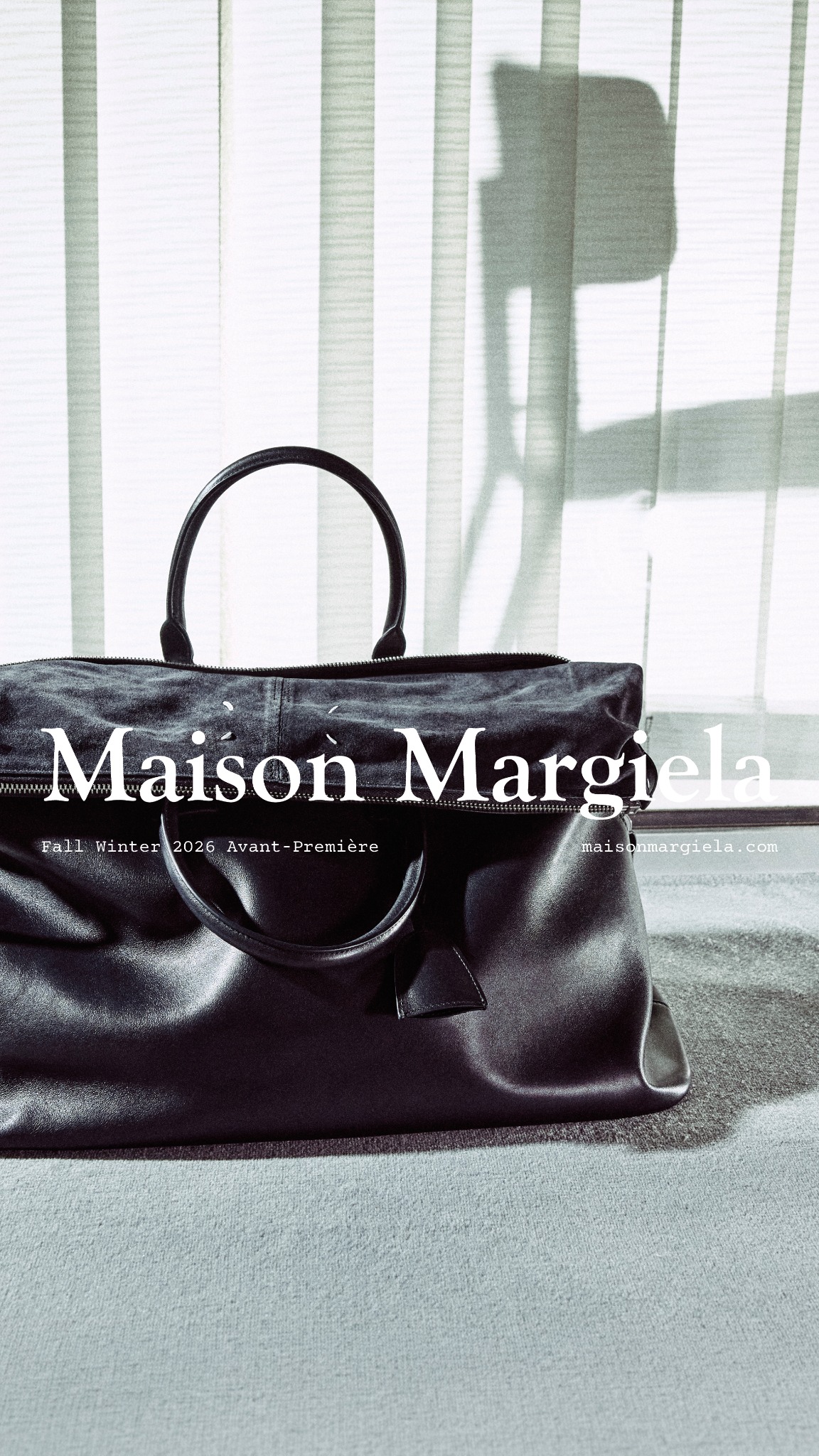 A close-up, low-angle shot of a dark leather Maison Margiela bag is presented against a backdrop of vertical blinds casting shadows. White text overlays the image, featuring the brand name "Maison Margiela" prominently. Below this, smaller text reads "Fall Winter 2026 Avant-Première" on the left and "maisonmargiela.com" on the right. The bag is the central focus, with its textured leather and handles clearly visible. The overall aesthetic is minimalist and high-fashion, highlighting the product's design and brand identity.