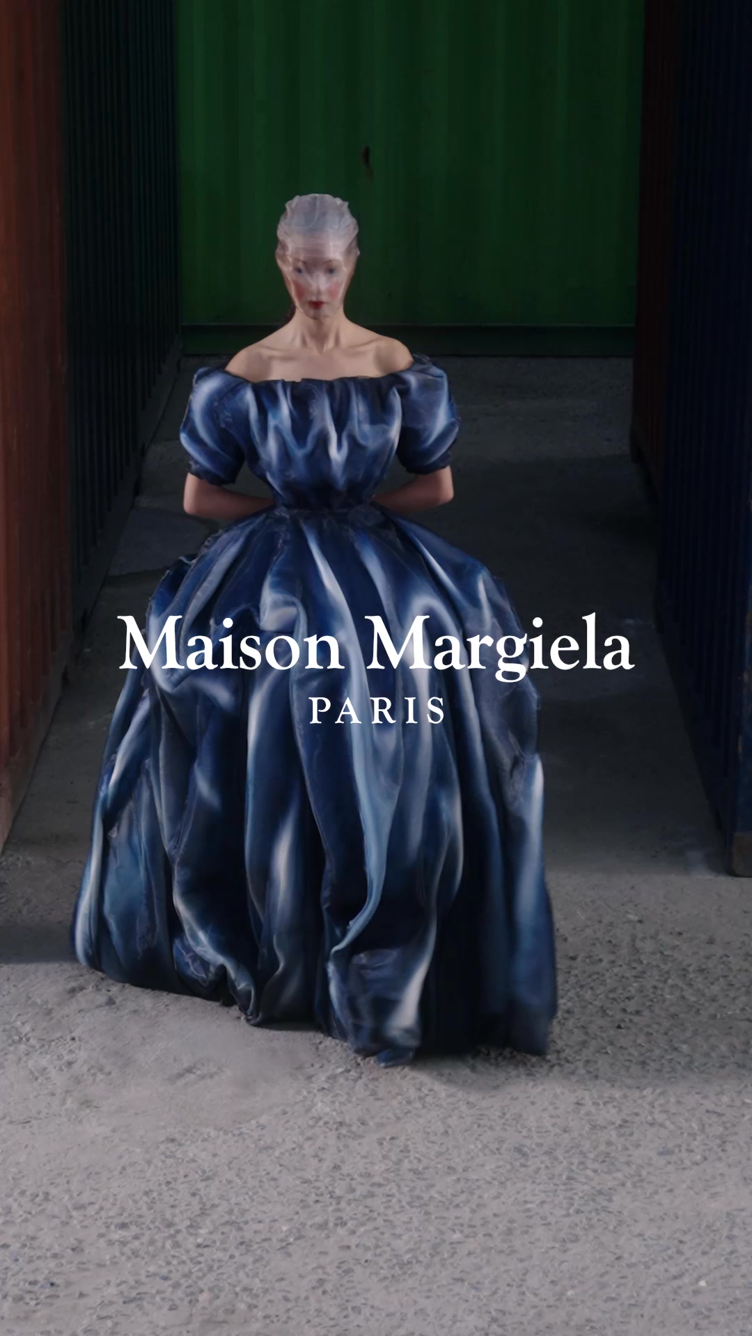 A model stands in a dramatic, voluminous blue and white marbled-patterned gown with off-the-shoulder sleeves. The background is industrial, with shipping containers visible. White text overlaid on the image reads "Maison Margiela" and below it, "PARIS". The copy invites the viewer to relive the Fall-Winter 2026 fashion show, explore looks, creation stages, and watch the full video, sparking curiosity about the collection.