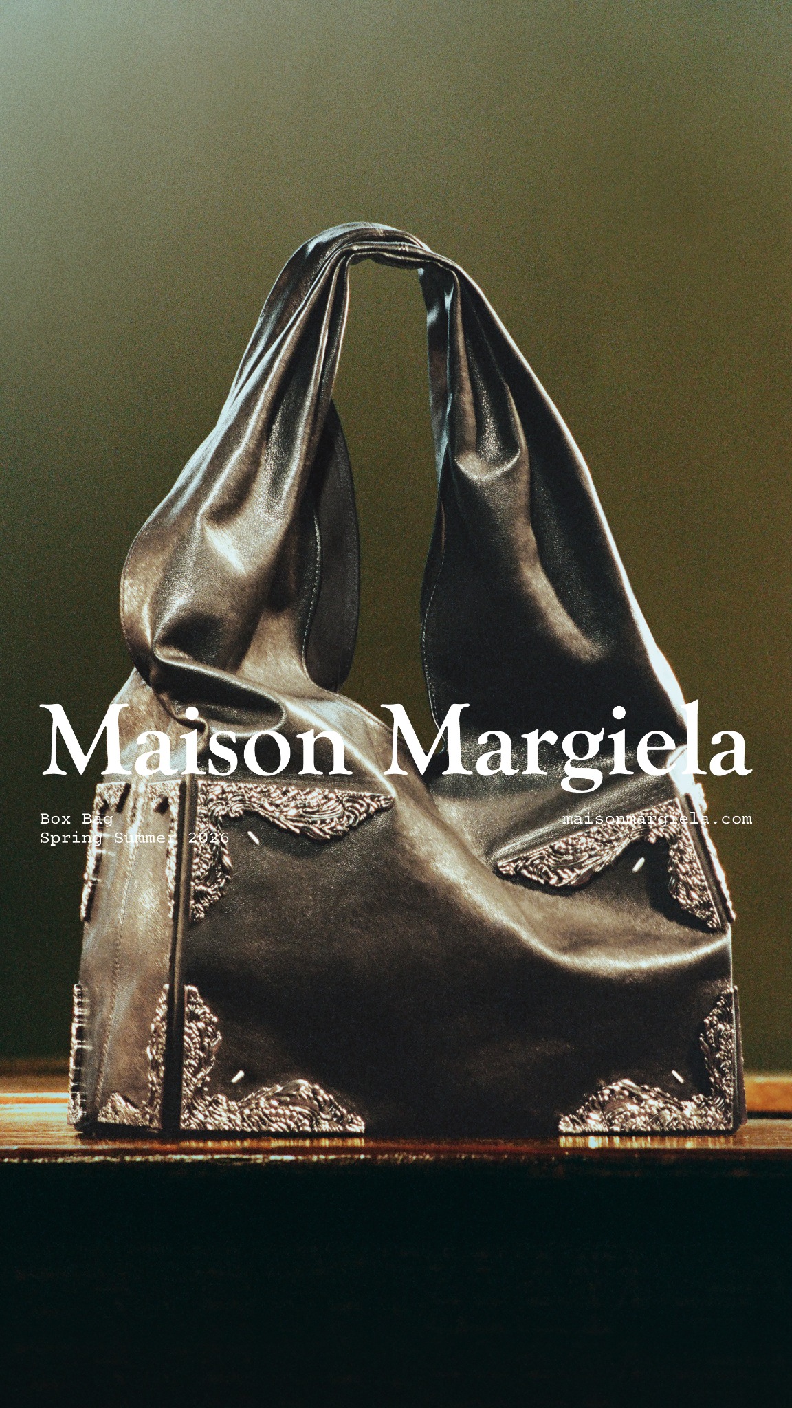 A dark, textured background frames a luxurious, black leather Maison Margiela handbag. The bag has a soft, draped silhouette and is adorned with ornate silver corner details. White text overlays the image, displaying the brand name "Maison Margiela" prominently, along with "Box Bag" and "Spring Summer 2026" in a smaller font. The website "maisonmargiela.com" is also visible.