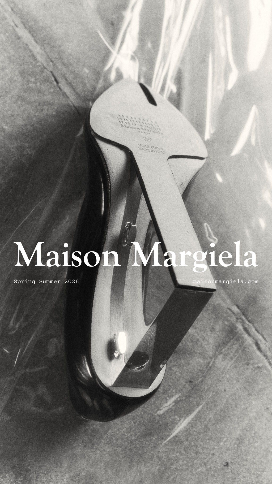 A black and white, close-up, overhead shot of a Maison Margiela shoe. The shoe has a split toe and a clear plexiglass heel. The brand name "Maison Margiela" is displayed prominently in large white serif font across the center of the image. Below it, in smaller white text, is "Spring Summer 2026" on the left and "maisonmargiela.com" on the right. The text overlay explains the shoe's design inspiration and materials.