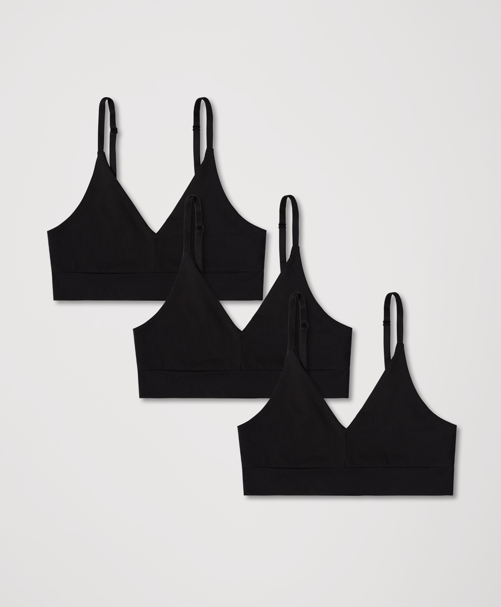 Three identical black bralettes are arranged in a staggered formation against a plain, light grey background. The bralettes are made of a soft, matte fabric and feature thin, adjustable straps and a V-neckline. The copy overlaying the image reads: "Made with organic cotton and designed to move with you — these are the pieces you’ll reach for again and again." The overall aesthetic is clean, minimalist, and focuses on the product's comfort and versatility.