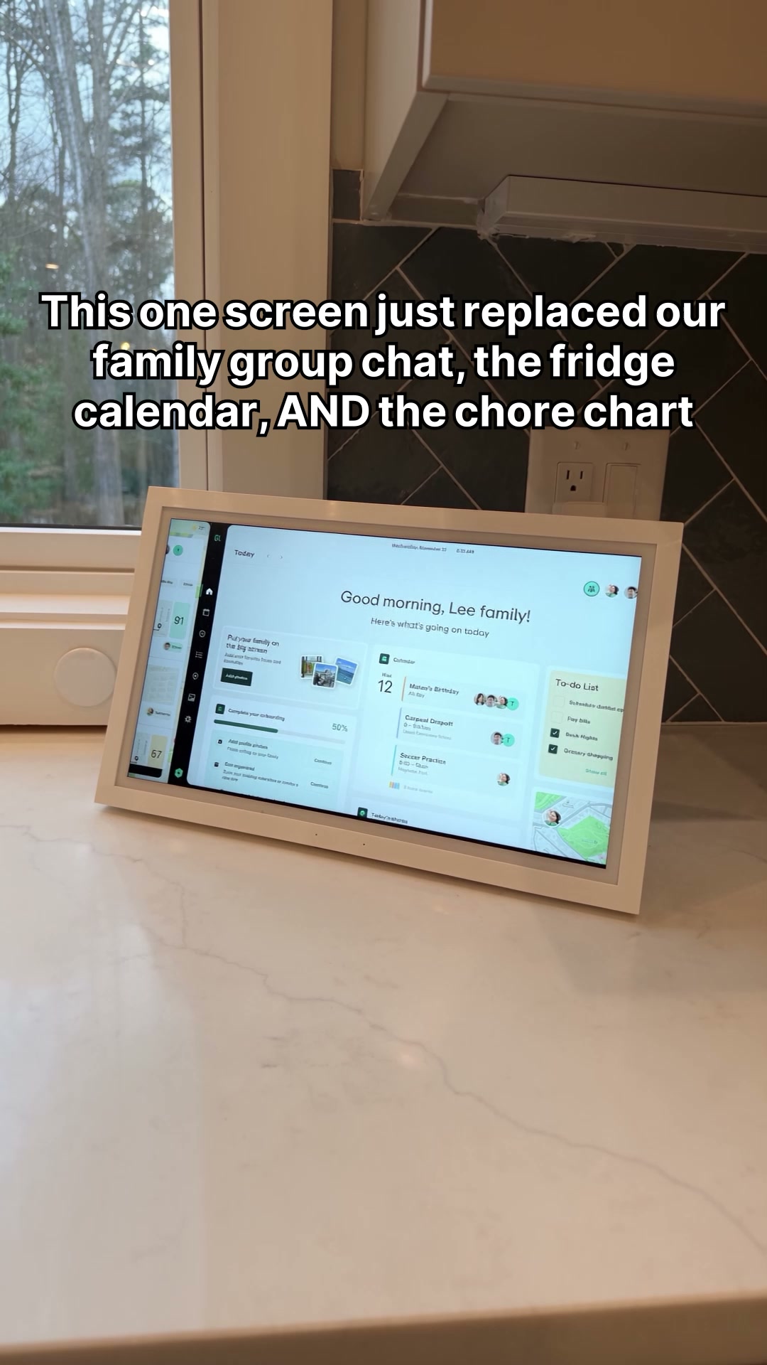 A digital smart display is shown on a kitchen counter. The screen displays a dashboard with various widgets, including a calendar, to-do list, and family photos. Large white text is overlaid on the image, stating "This one screen just replaced our family group chat, the fridge calendar, AND the chore chart." The display appears to be a central hub for family organization.